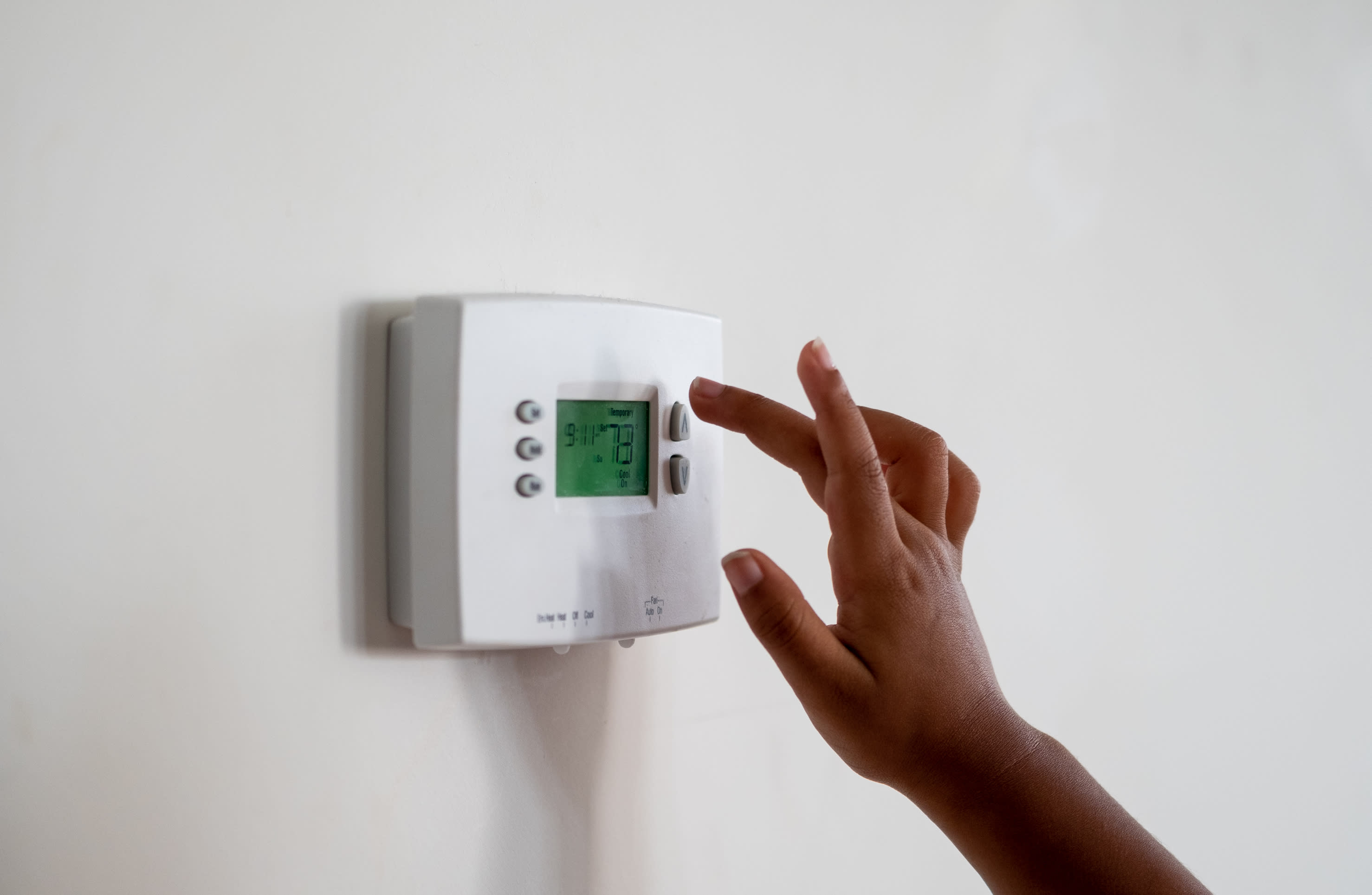 A white thermostat mounted on a wall, with a hand adjusting the temperature setting on the digital display.