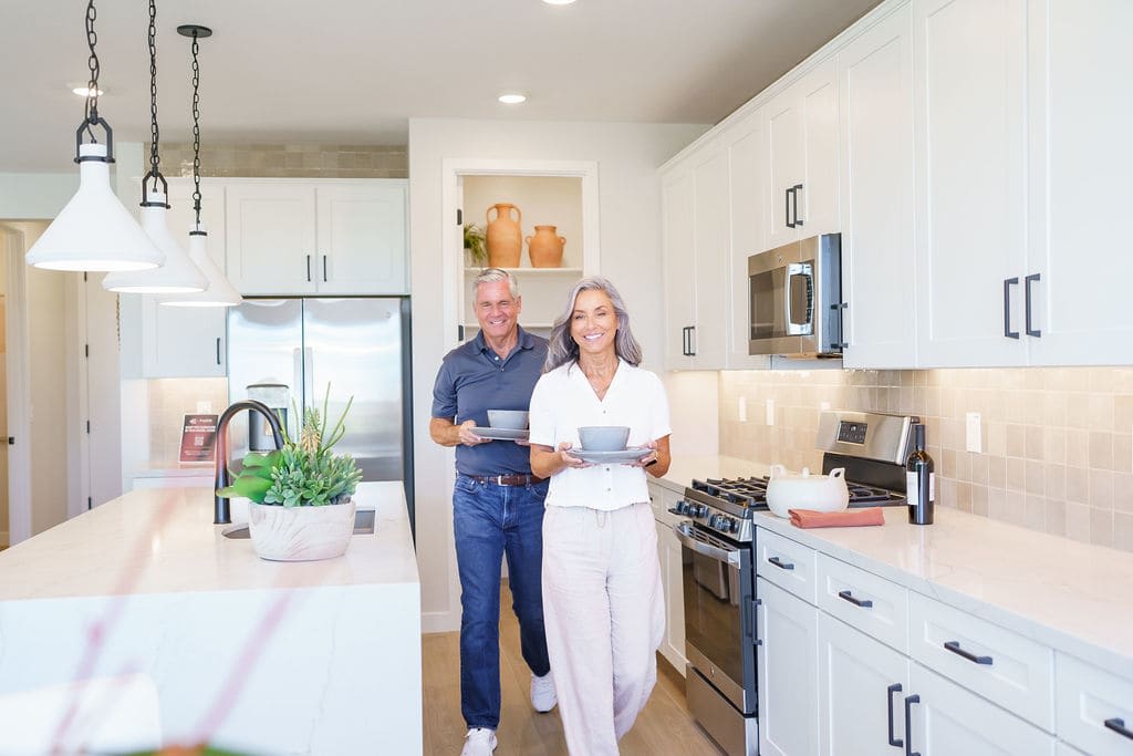 A middle-aged couple stands in a modern, well-equipped kitchen, with the woman holding a cup and the man standing beside her.