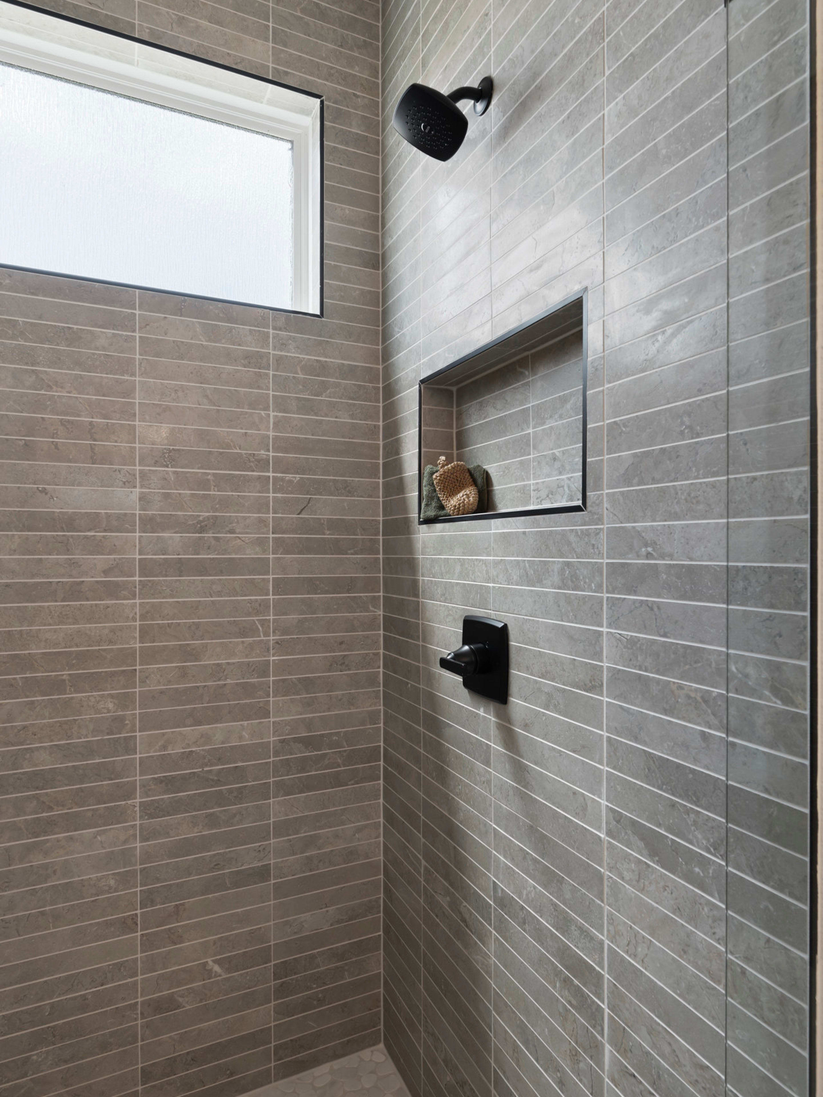 A modern, tiled shower enclosure with a recessed shelf and a mounted showerhead, set against a backdrop of a large window providing natural lighting.