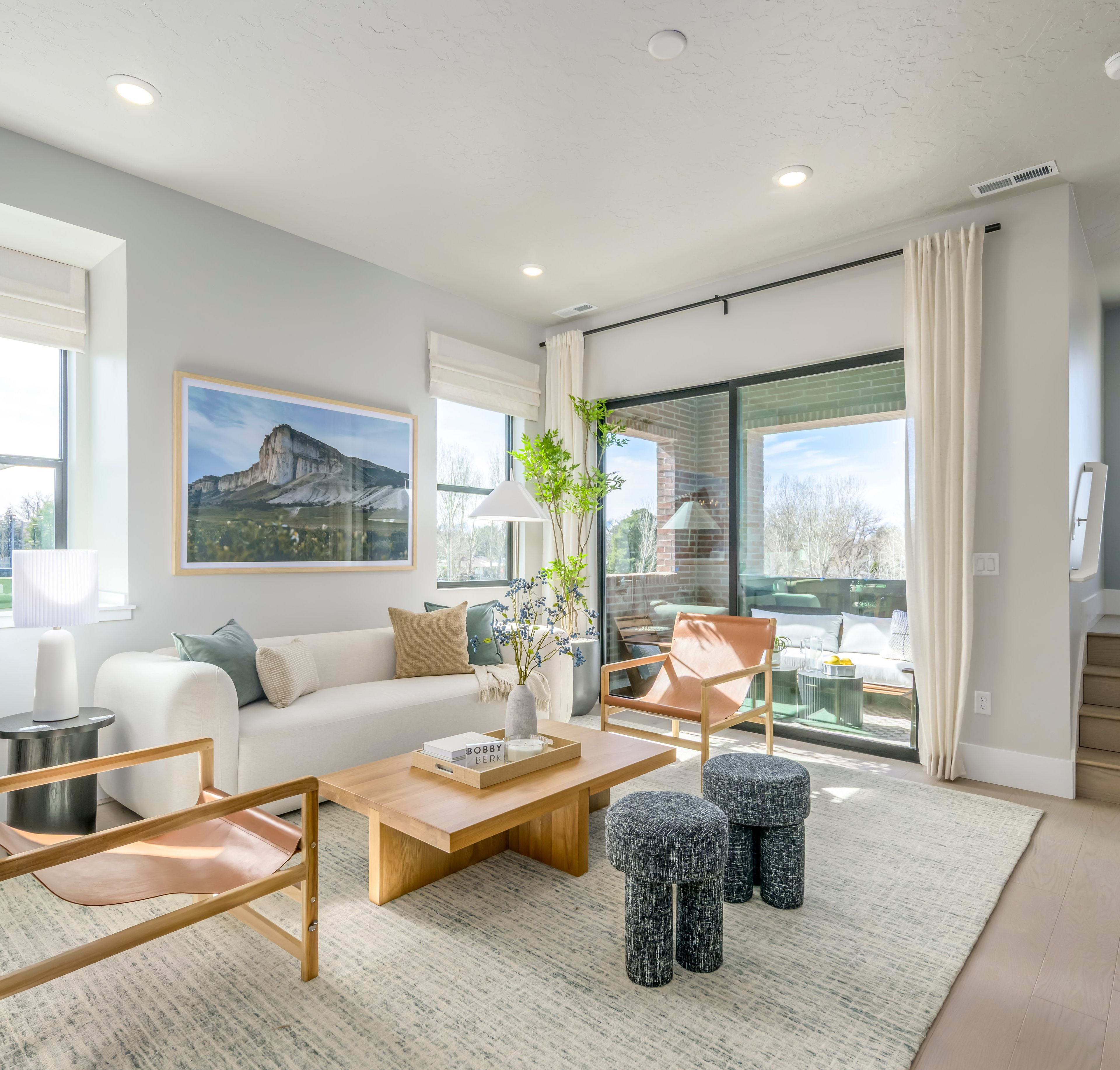 A cozy and modern living room with large windows offering a scenic view of a mountainous landscape, featuring a white sofa, wooden furniture, and decorative elements that create a warm and inviting atmosphere.