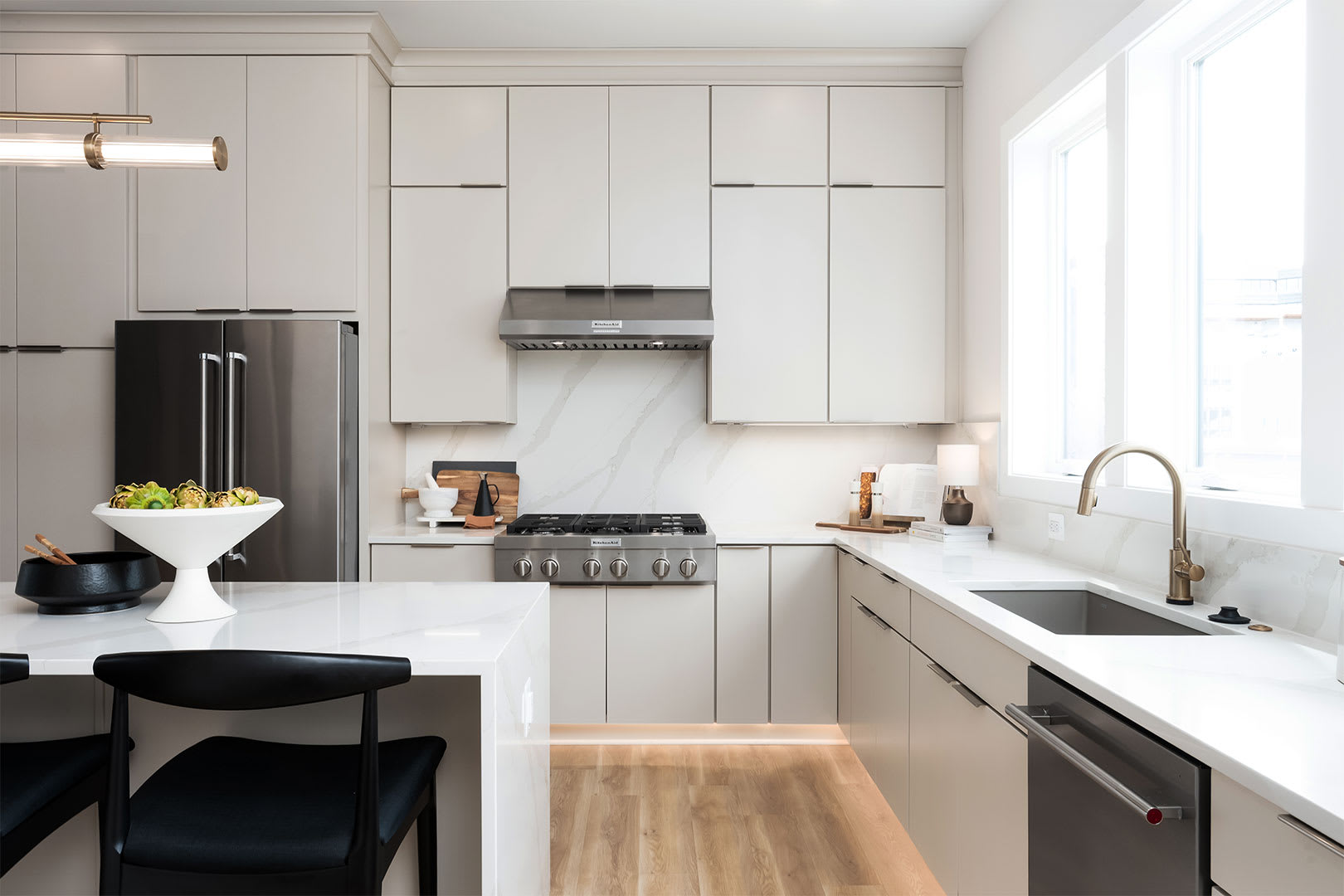A modern, minimalist kitchen with white cabinets, stainless steel appliances, and a wooden floor, featuring a dining area with black chairs.
