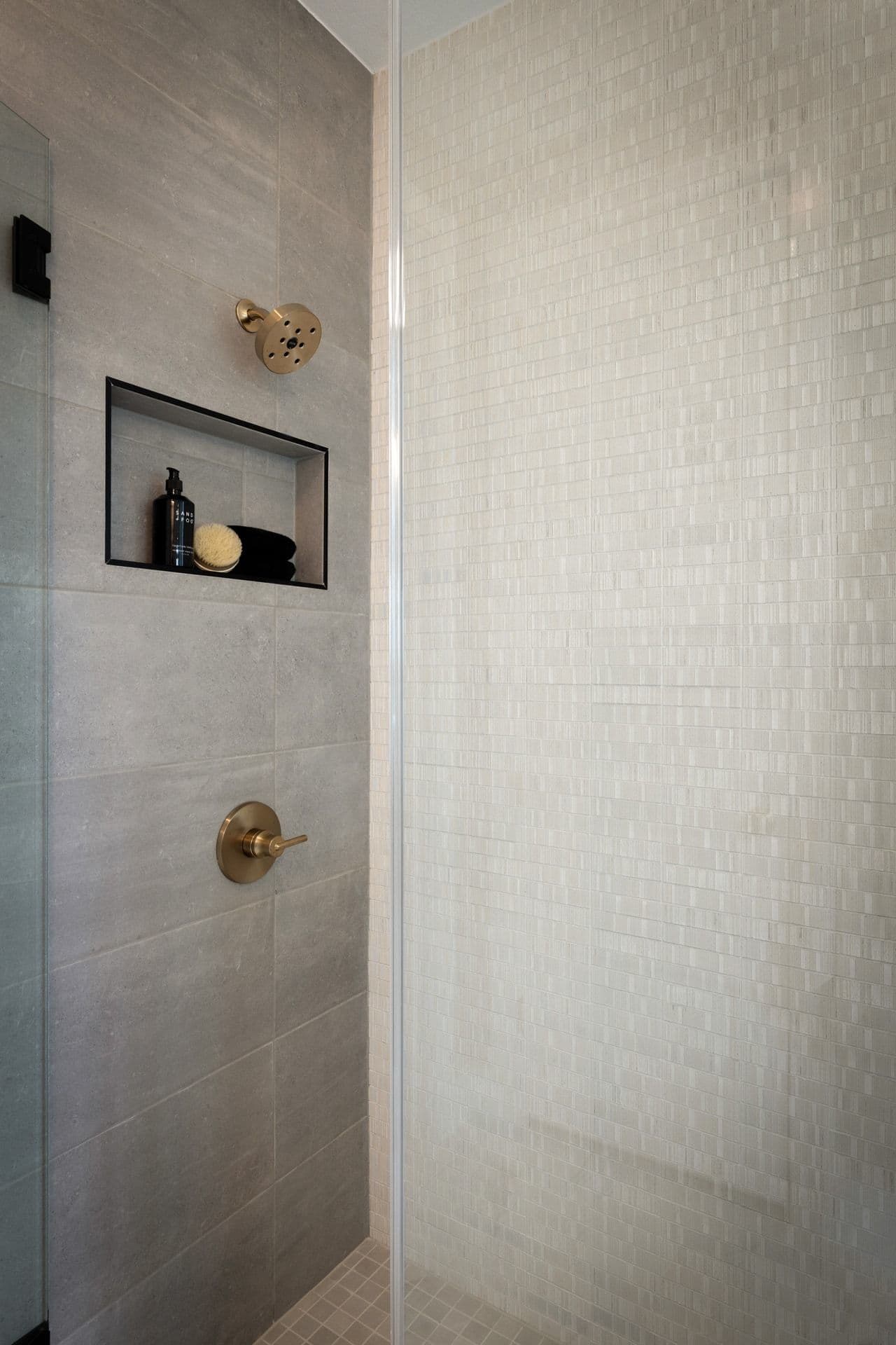 A modern shower stall features a built-in niche with toiletries in the foreground, and a textured tiled wall in the background.