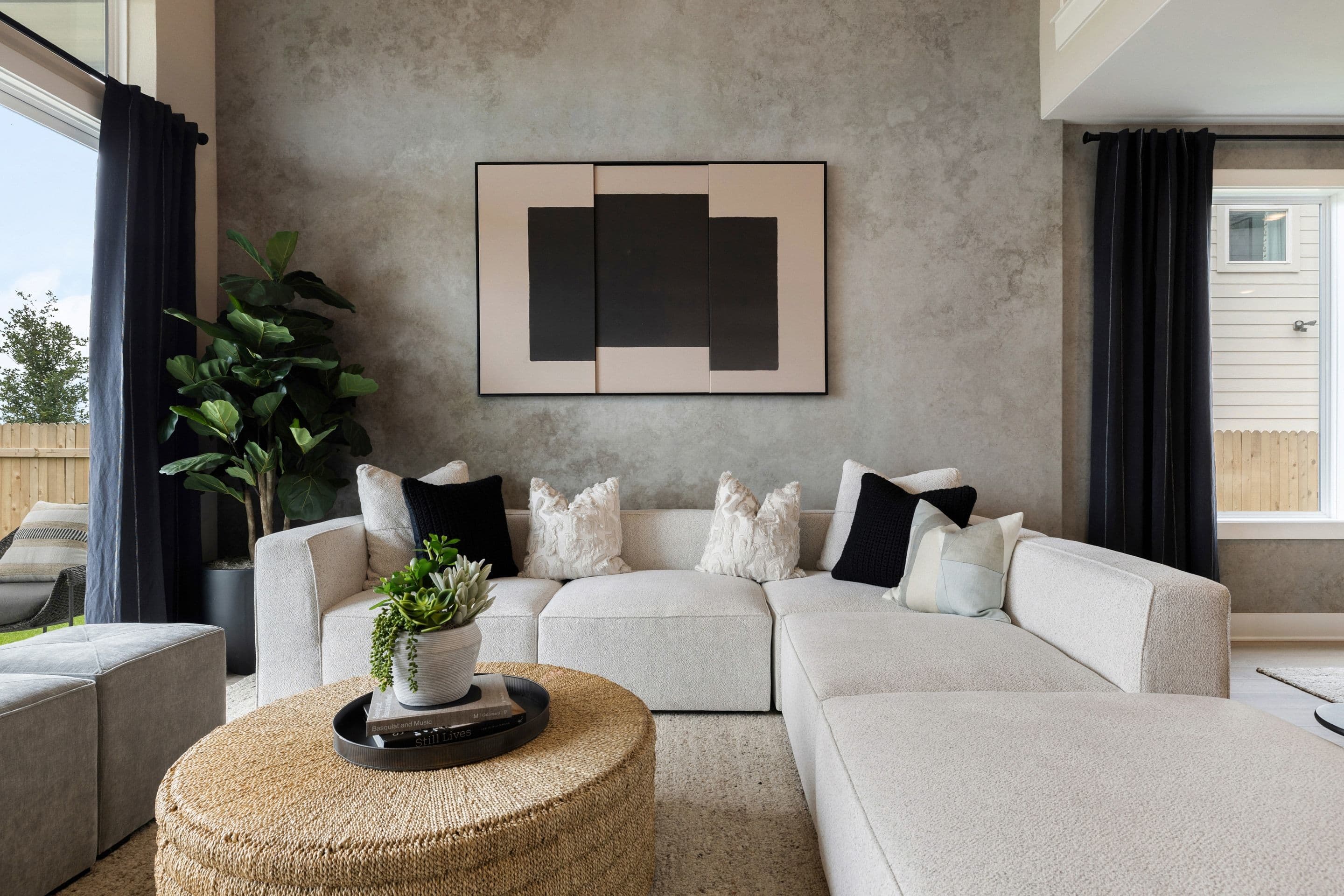 A large sectional sofa with decorative pillows sits in front of a textured gray wall with abstract art, while a round, woven ottoman and a potted plant are in the foreground.