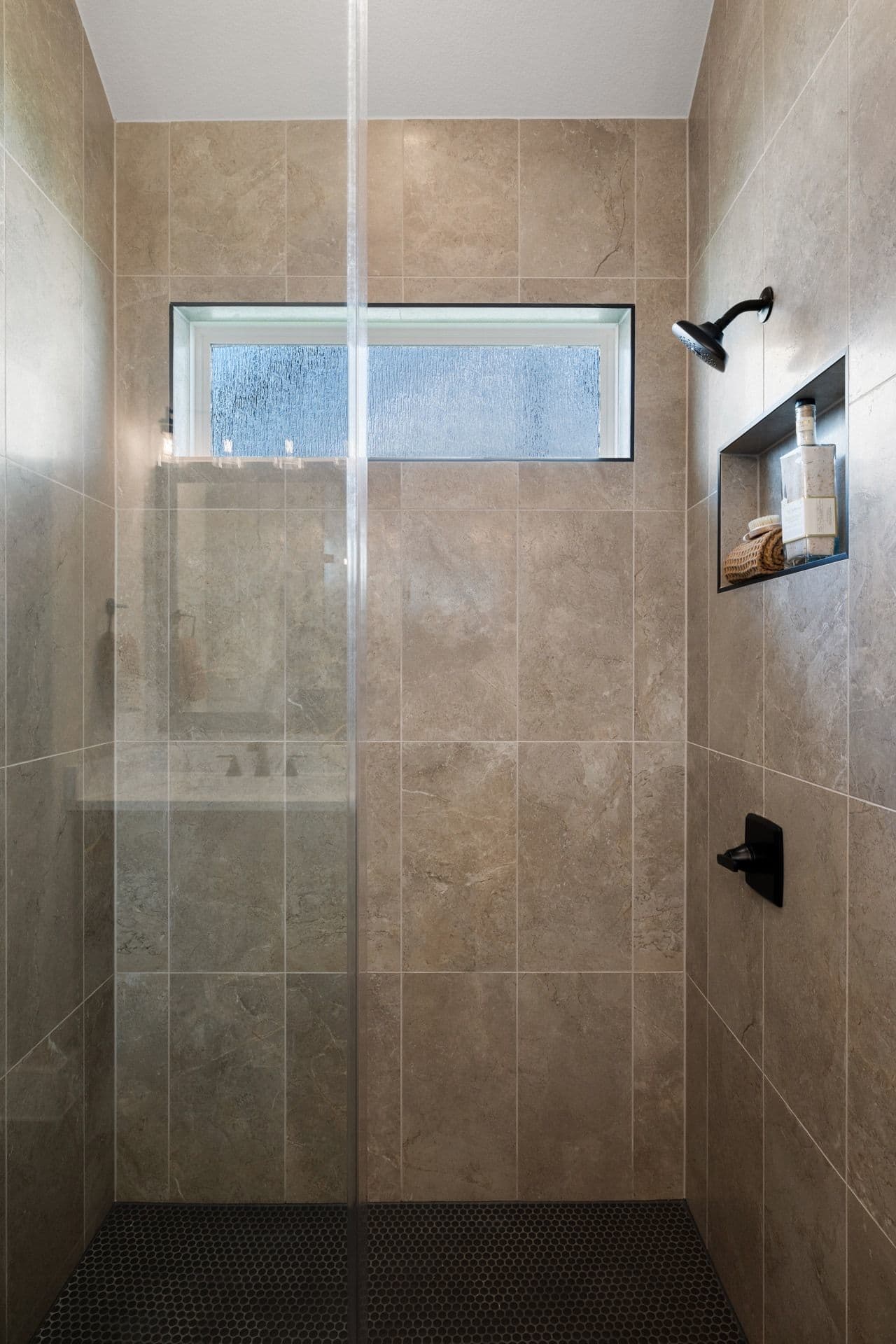 A modern shower stall with a glass door in the foreground and tiled walls with a small window and showerhead in the background.