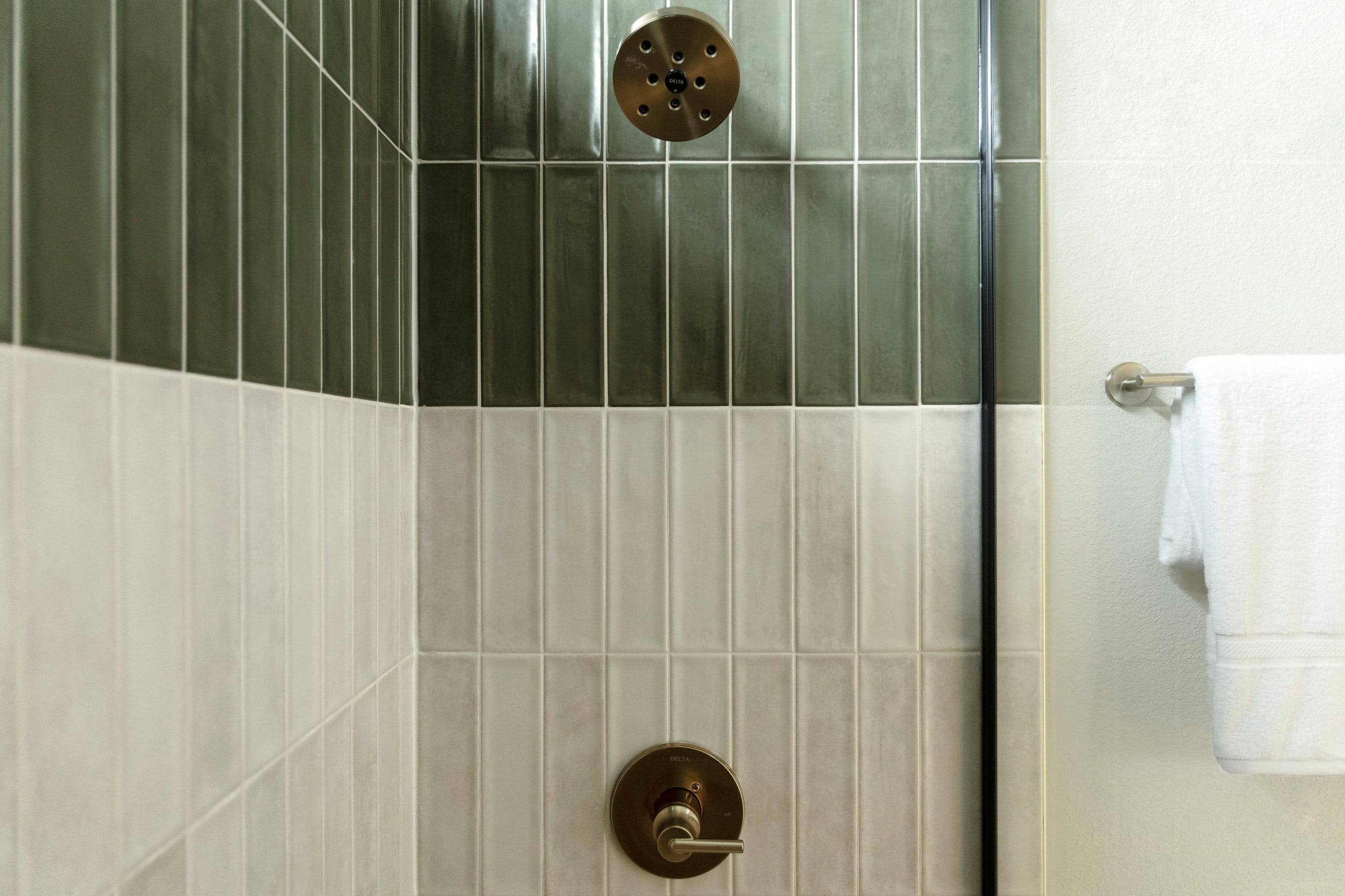 A modern shower stall features a brass showerhead and handle in the foreground, with a tiled wall and a white towel hanging on a rack in the background.