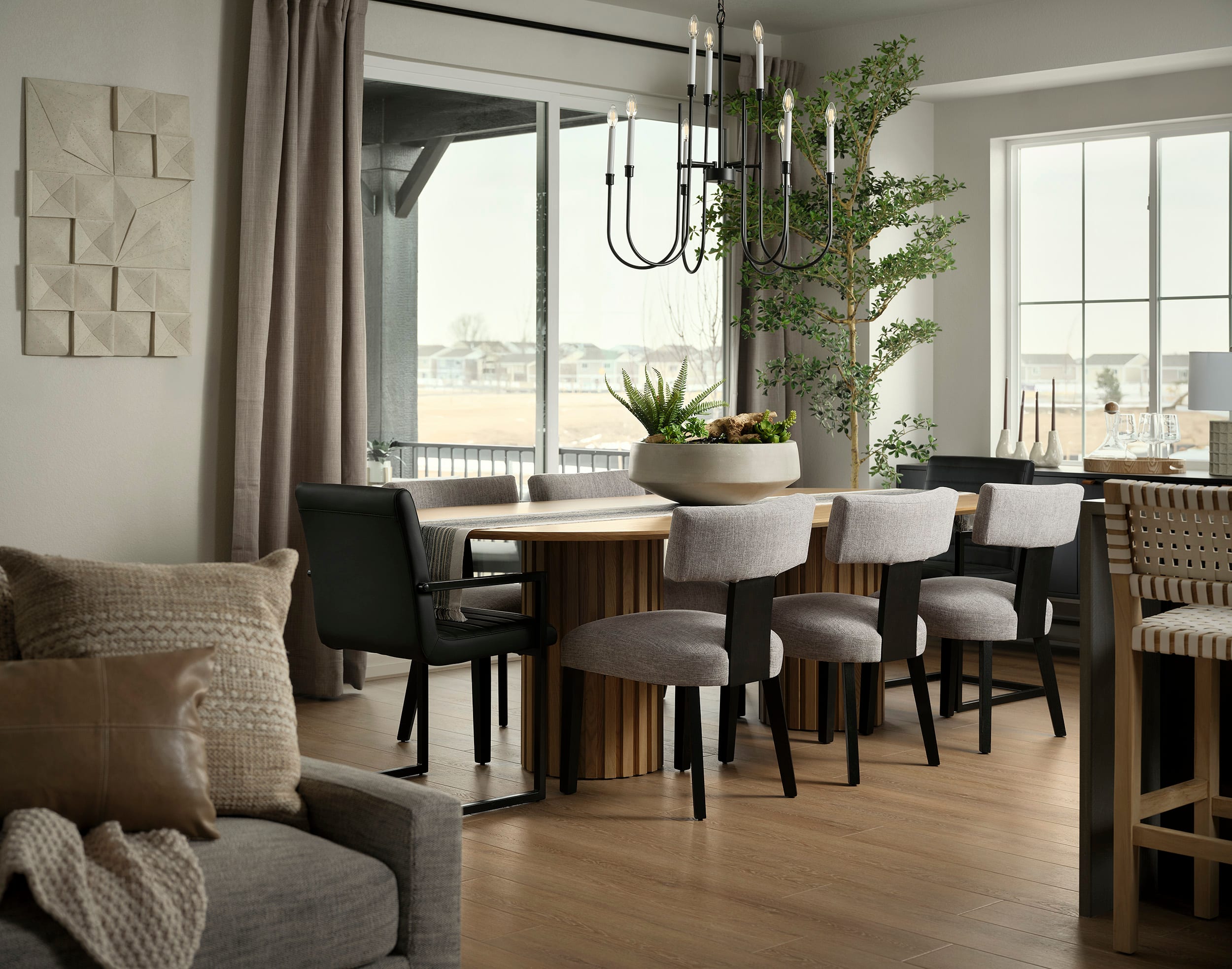 A cozy and modern dining room with a large window, a wooden table surrounded by upholstered chairs, and a decorative chandelier hanging above, complemented by potted plants and textured wall art.