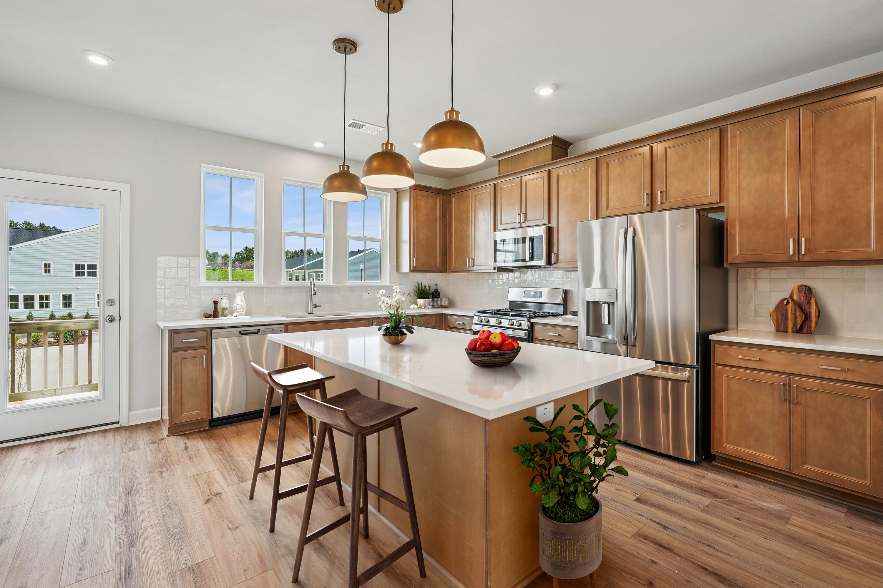 A modern and spacious kitchen with wooden cabinets, stainless steel appliances, a central island, and pendant lighting fixtures, set against a backdrop of large windows overlooking a scenic outdoor view.
