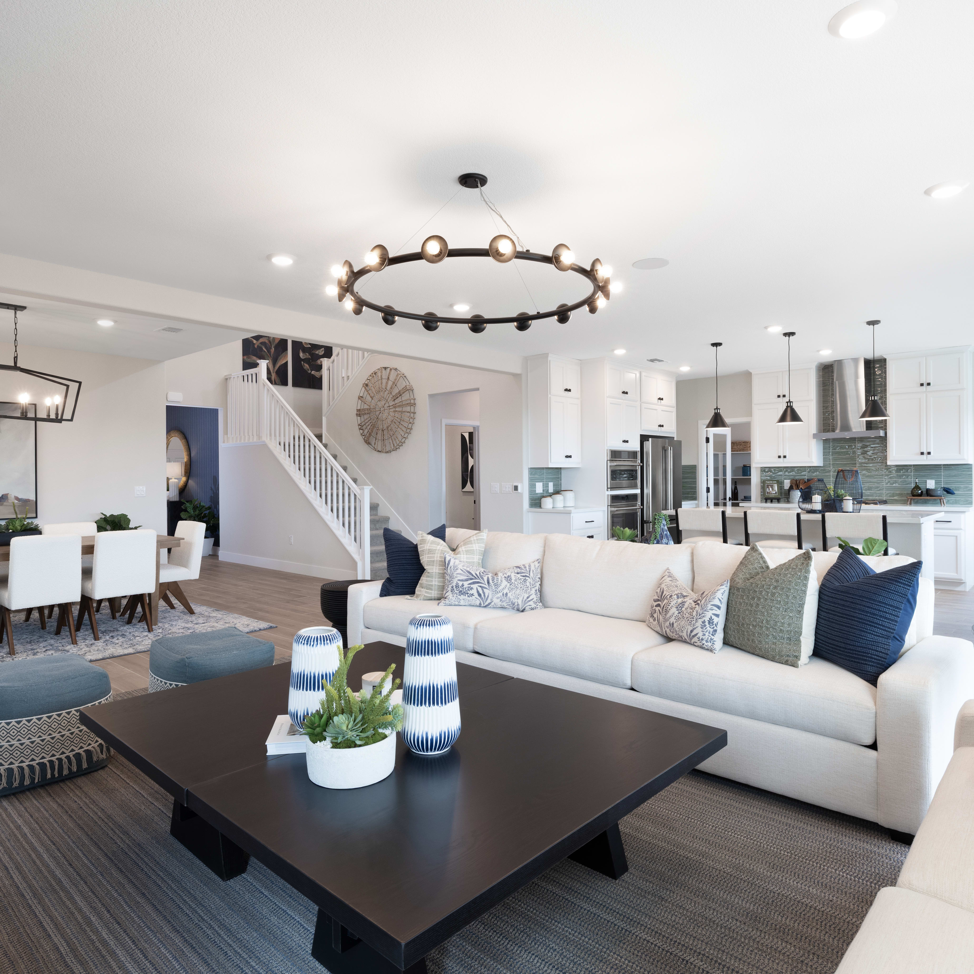 A spacious and well-designed living room with a large sofa, coffee table, and a chandelier overhead, surrounded by a dining area and an open-concept kitchen in the background.