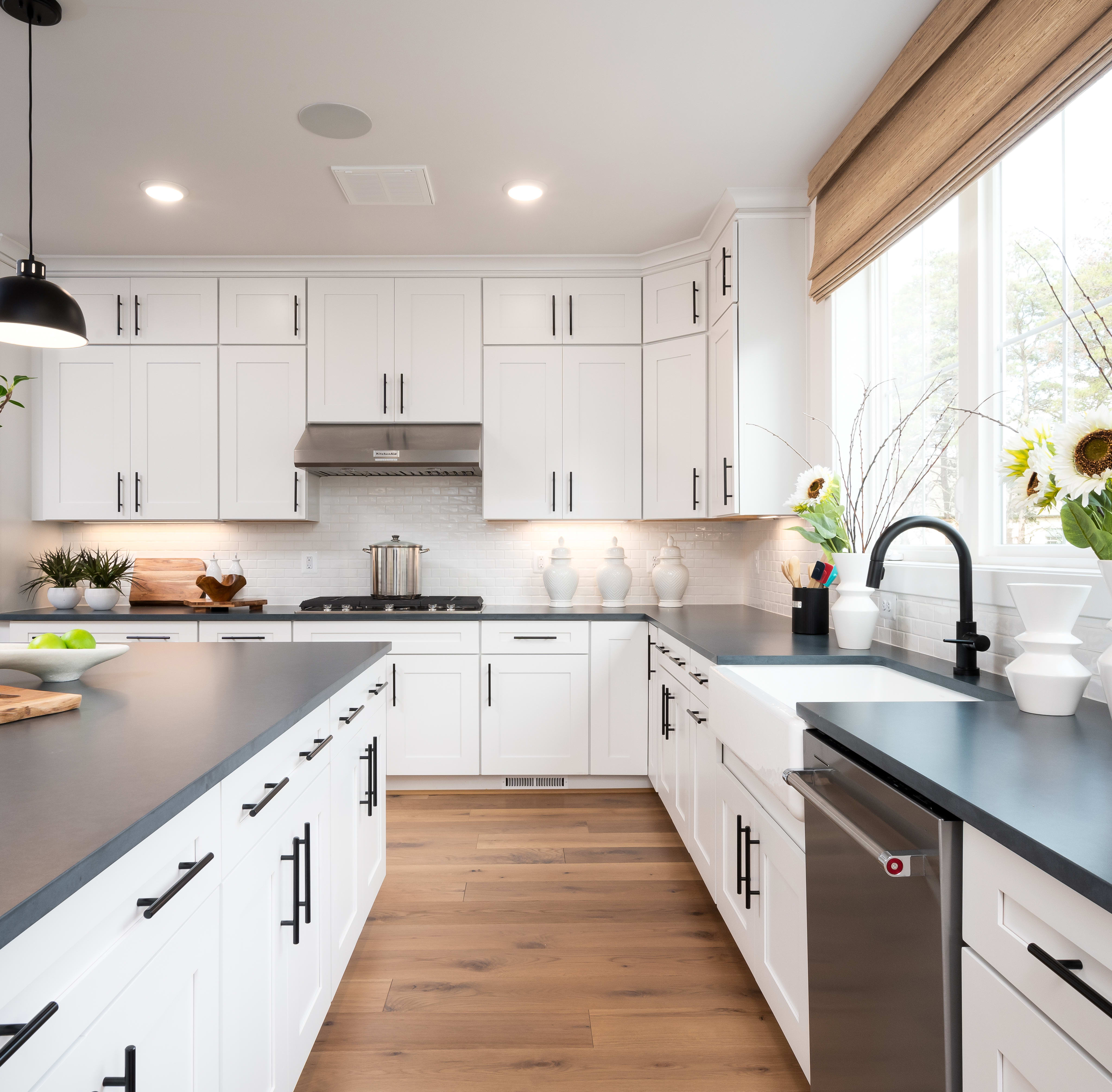 A modern, bright kitchen with white cabinets, gray countertops, and wooden floors, featuring a large window that lets in natural light and showcases a vase of sunflowers on the windowsill.