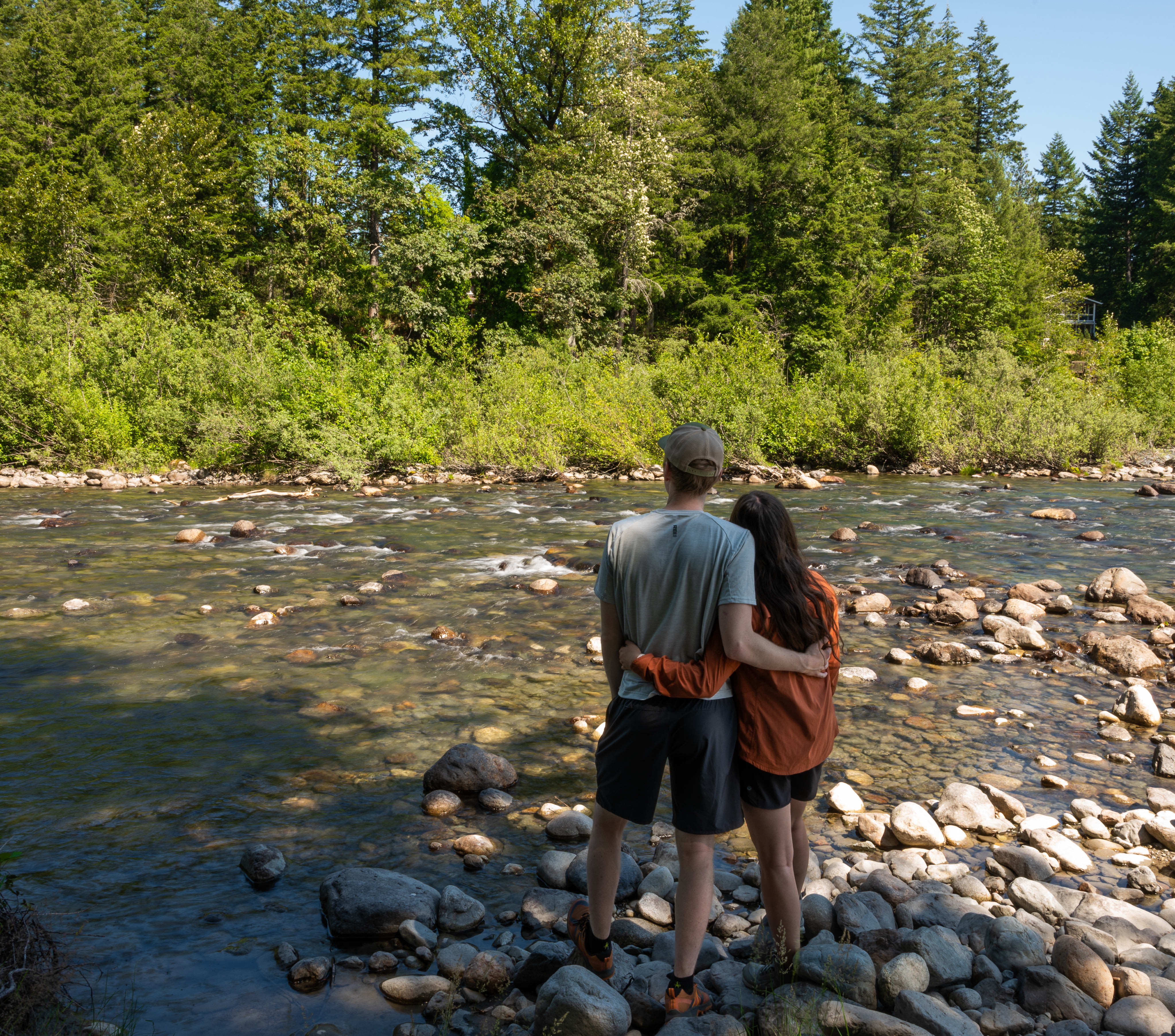 Two people standing on the rocky shore of a river, surrounded by a lush, forested landscape.