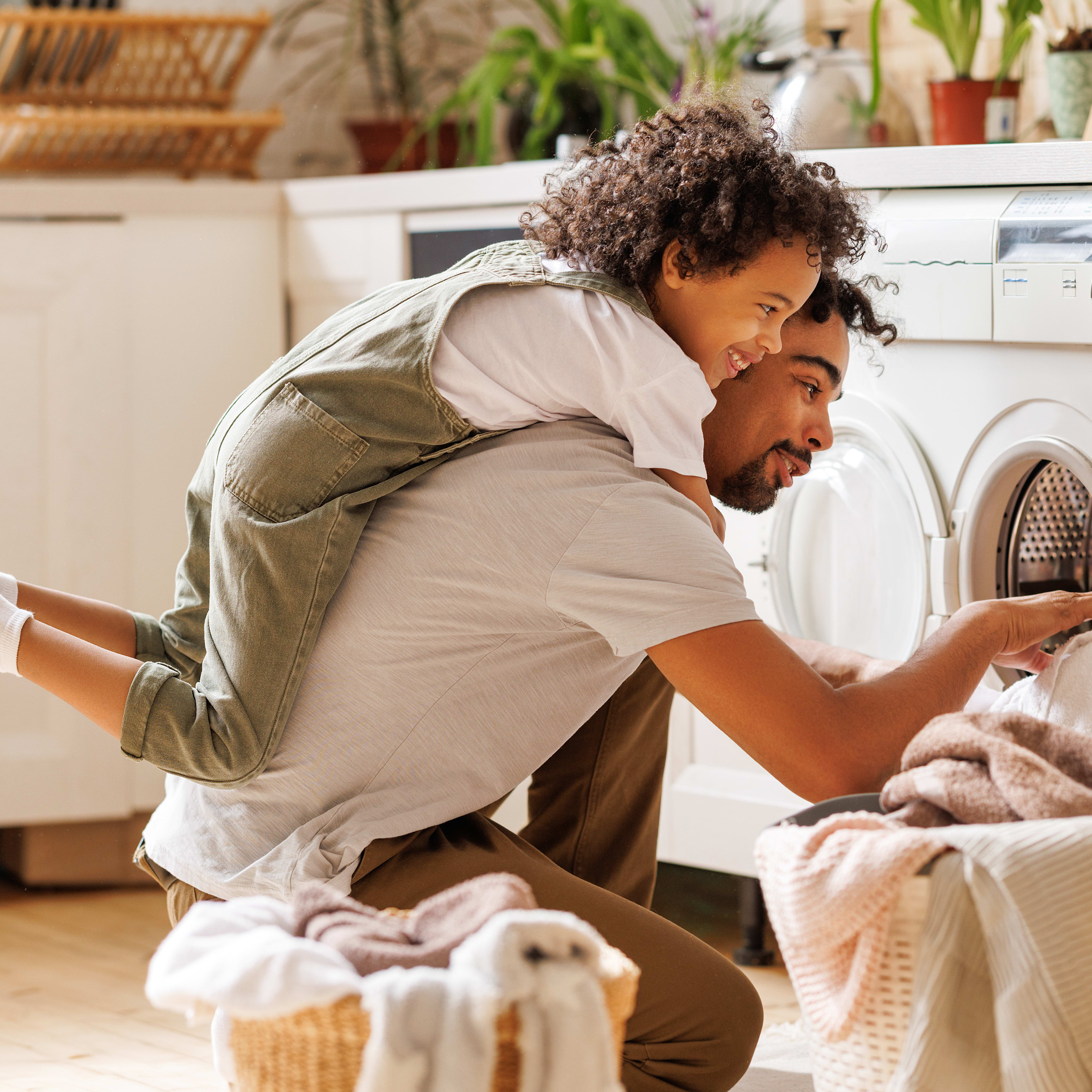 A man with curly hair is crouched down in front of a washing machine, sorting through a pile of laundry in a cozy, plant-filled home environment.