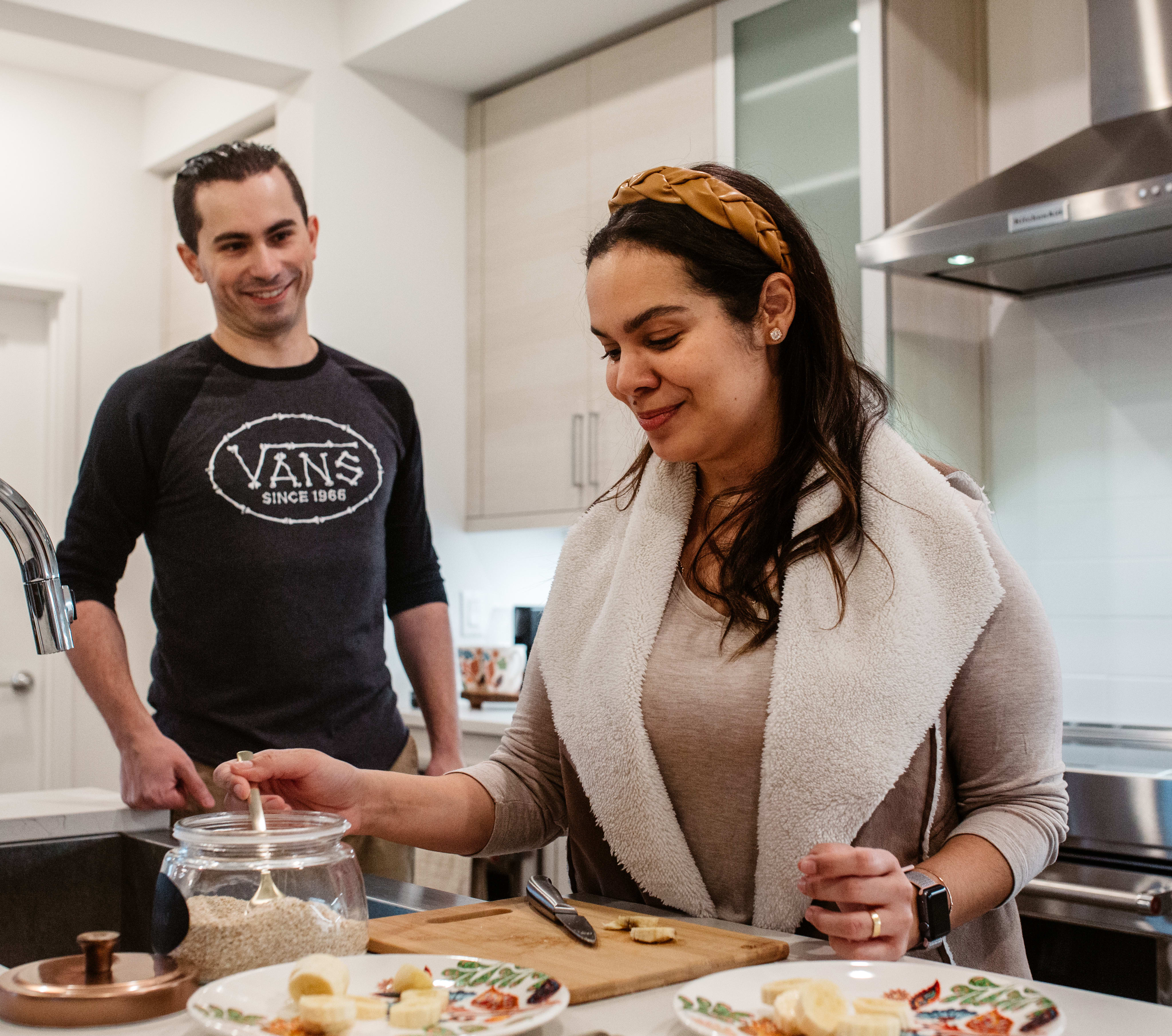 A young couple is preparing food together in a modern kitchen, with the woman wearing a headband and the man wearing a Vans t-shirt.