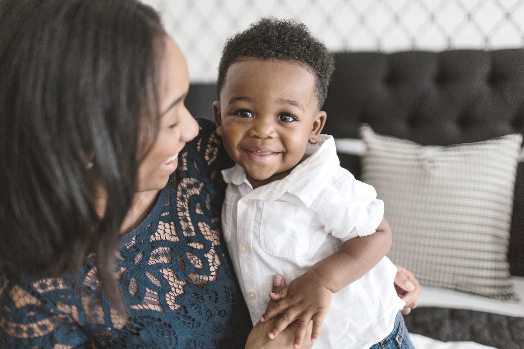 A woman in a patterned dress is holding a smiling young boy with curly hair in her arms, both of them sitting on a couch with decorative pillows in the background.