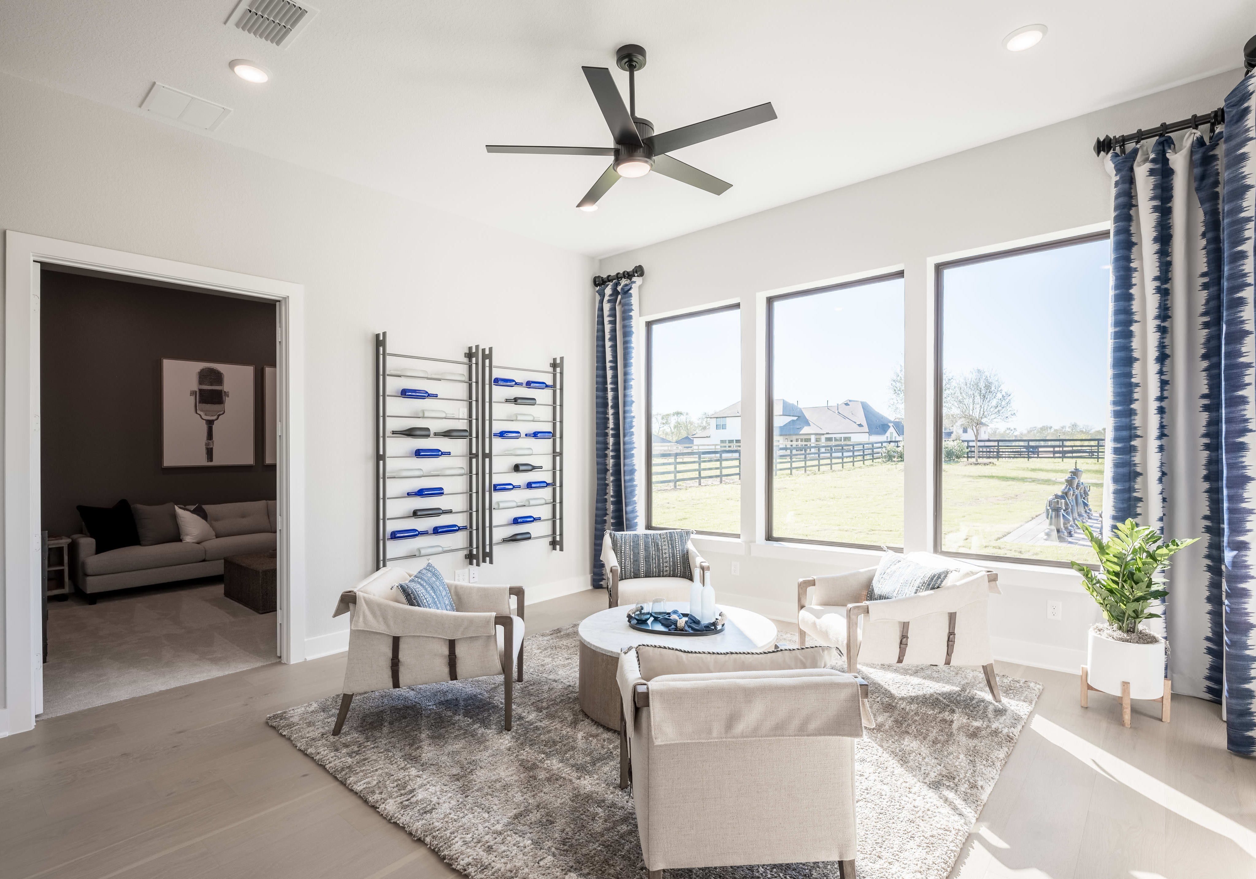 A spacious and well-lit living room with a ceiling fan, large windows overlooking a scenic outdoor area, and comfortable seating arrangements including armchairs and a sofa.