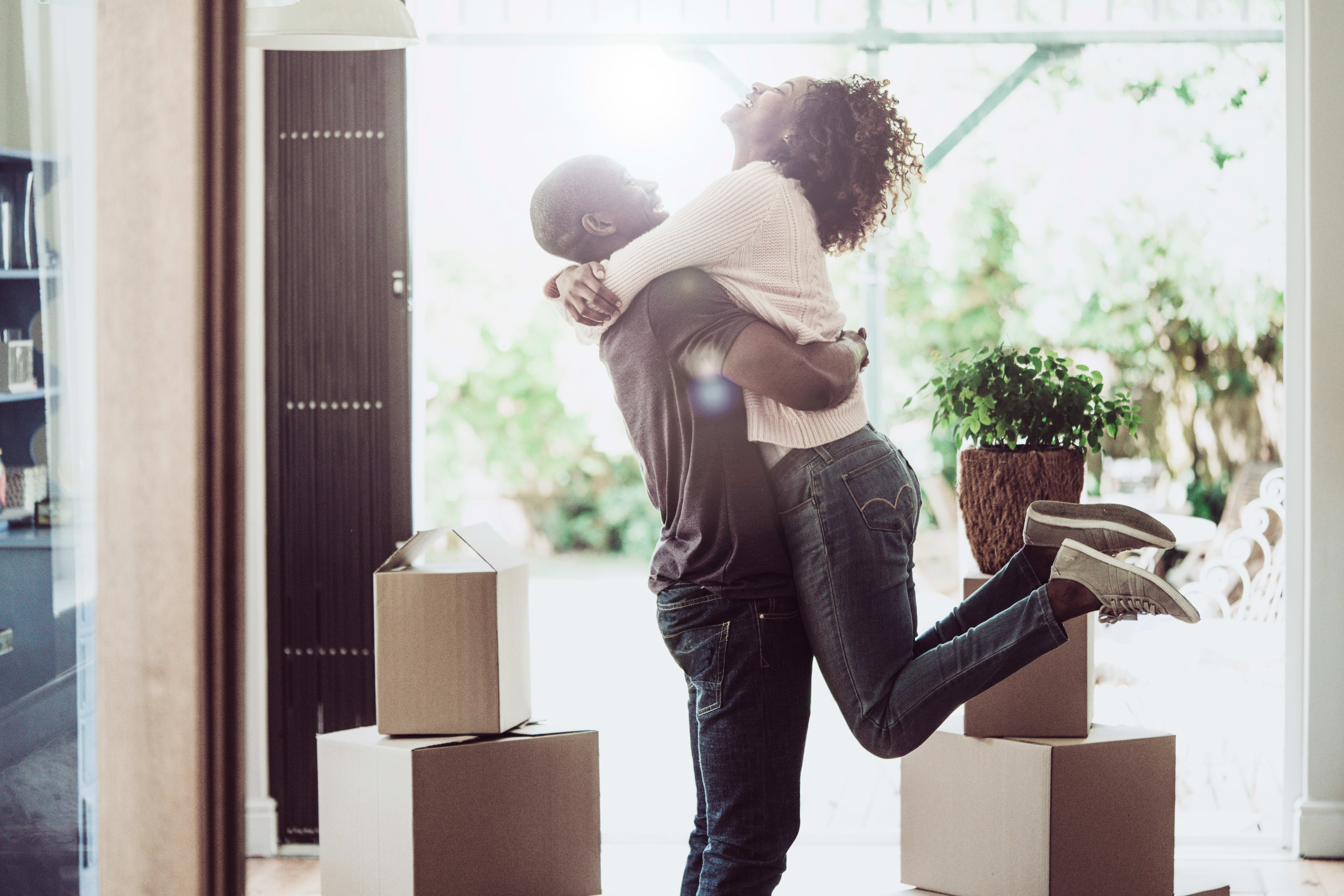 A couple embracing in a room surrounded by moving boxes, with a window in the background providing natural lighting.