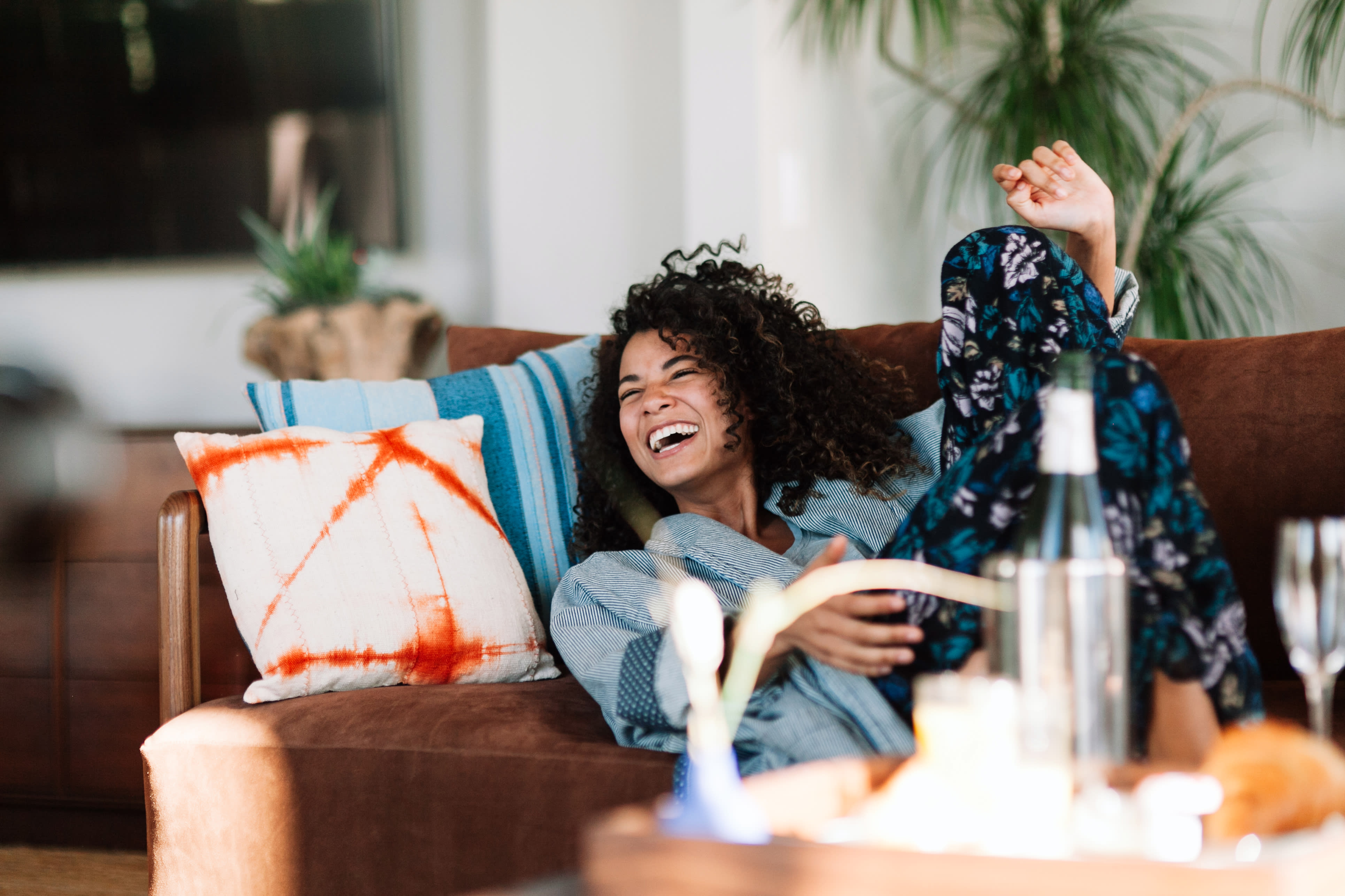 A young woman with curly hair sits on a couch, laughing joyfully while surrounded by colorful pillows and plants in the background.