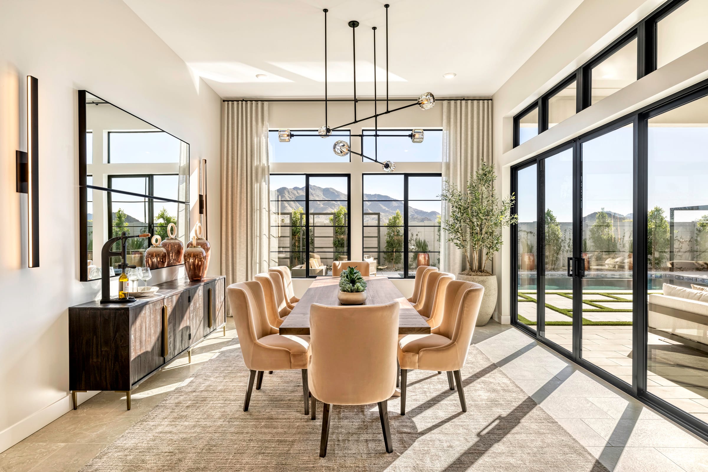 A spacious and modern dining room with large windows overlooking a scenic outdoor area, featuring a wooden dining table surrounded by plush beige chairs and accented by a sleek black chandelier.