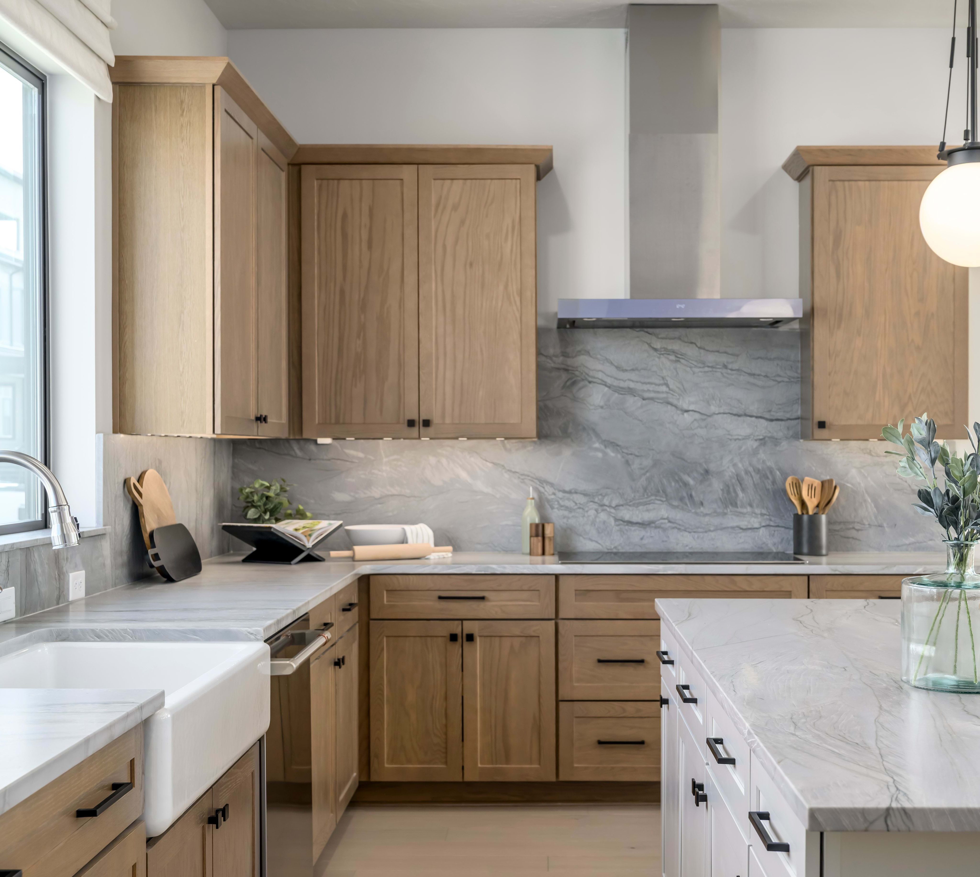 A modern, well-designed kitchen with wooden cabinets, a large marble countertop, and a farmhouse-style sink in the foreground, set against a backdrop of large windows providing ample natural light.