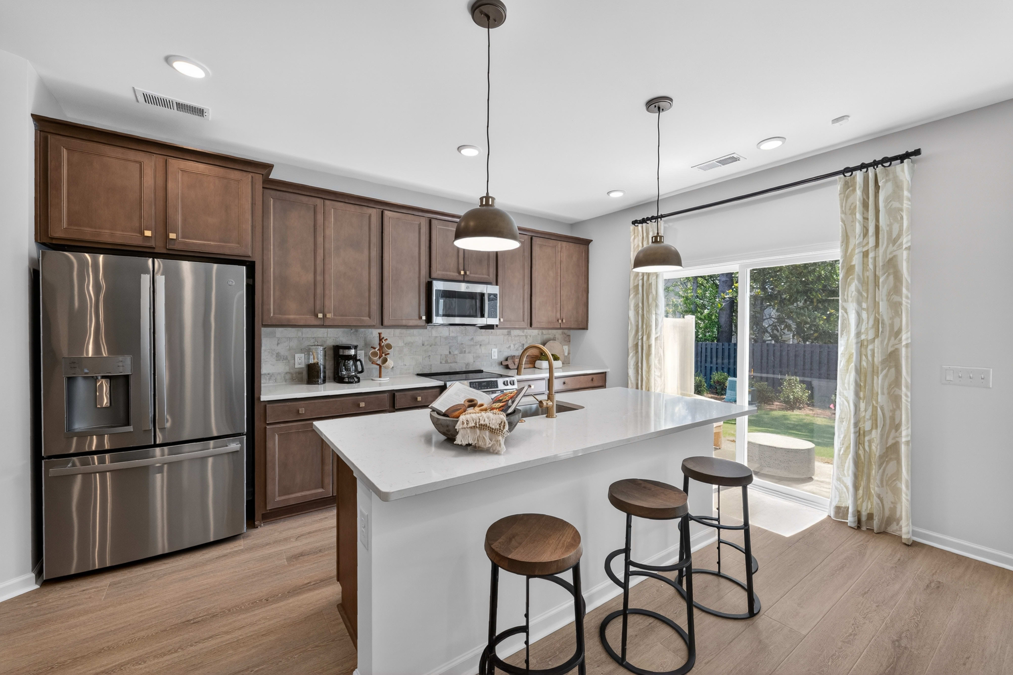 A modern kitchen with a stainless steel refrigerator and island with bar stools in the foreground, and a sliding glass door leading to a backyard in the background.