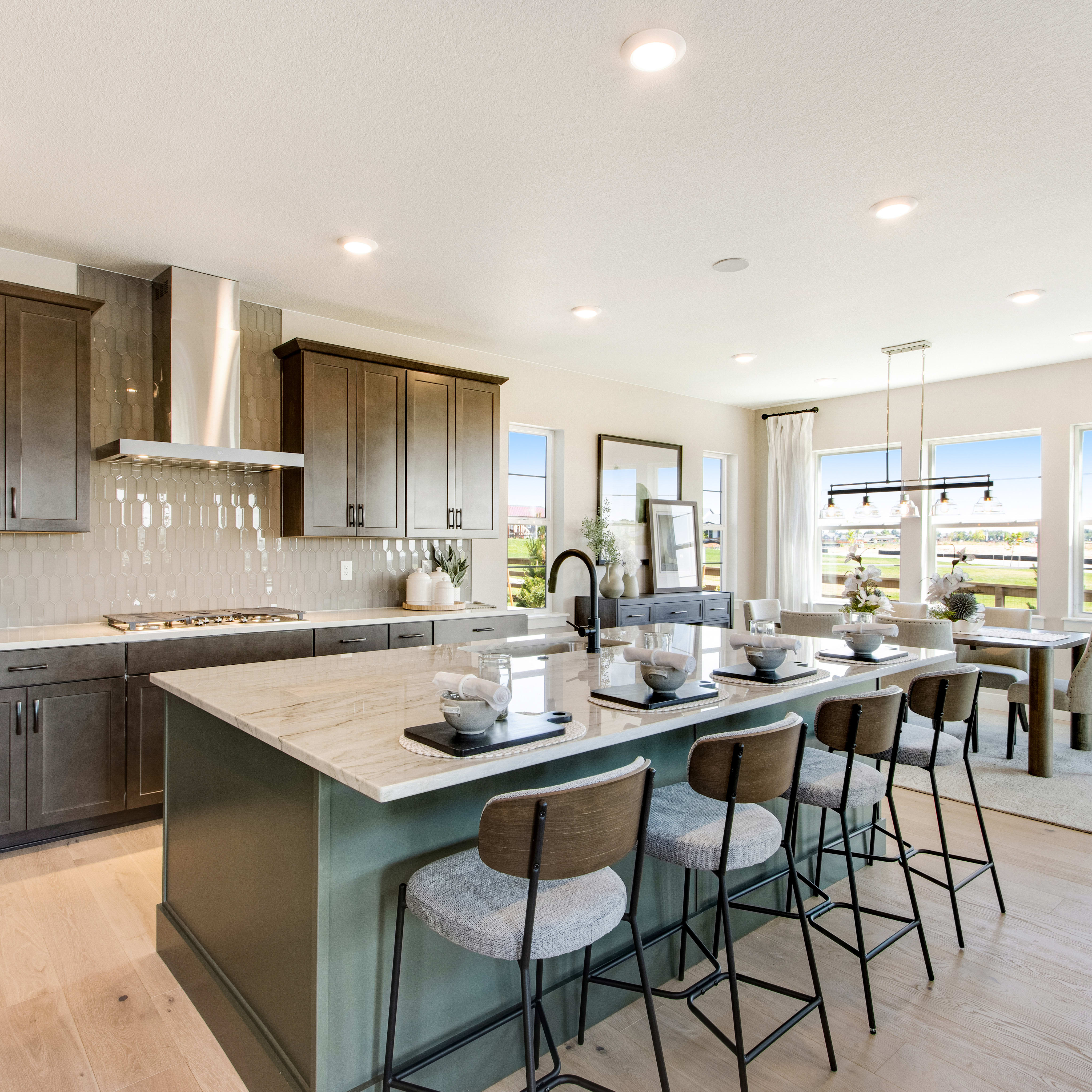 A modern kitchen island with bar stools in the foreground faces a dining area with a table and chairs, all set against a backdrop of large windows overlooking a sunny landscape.
