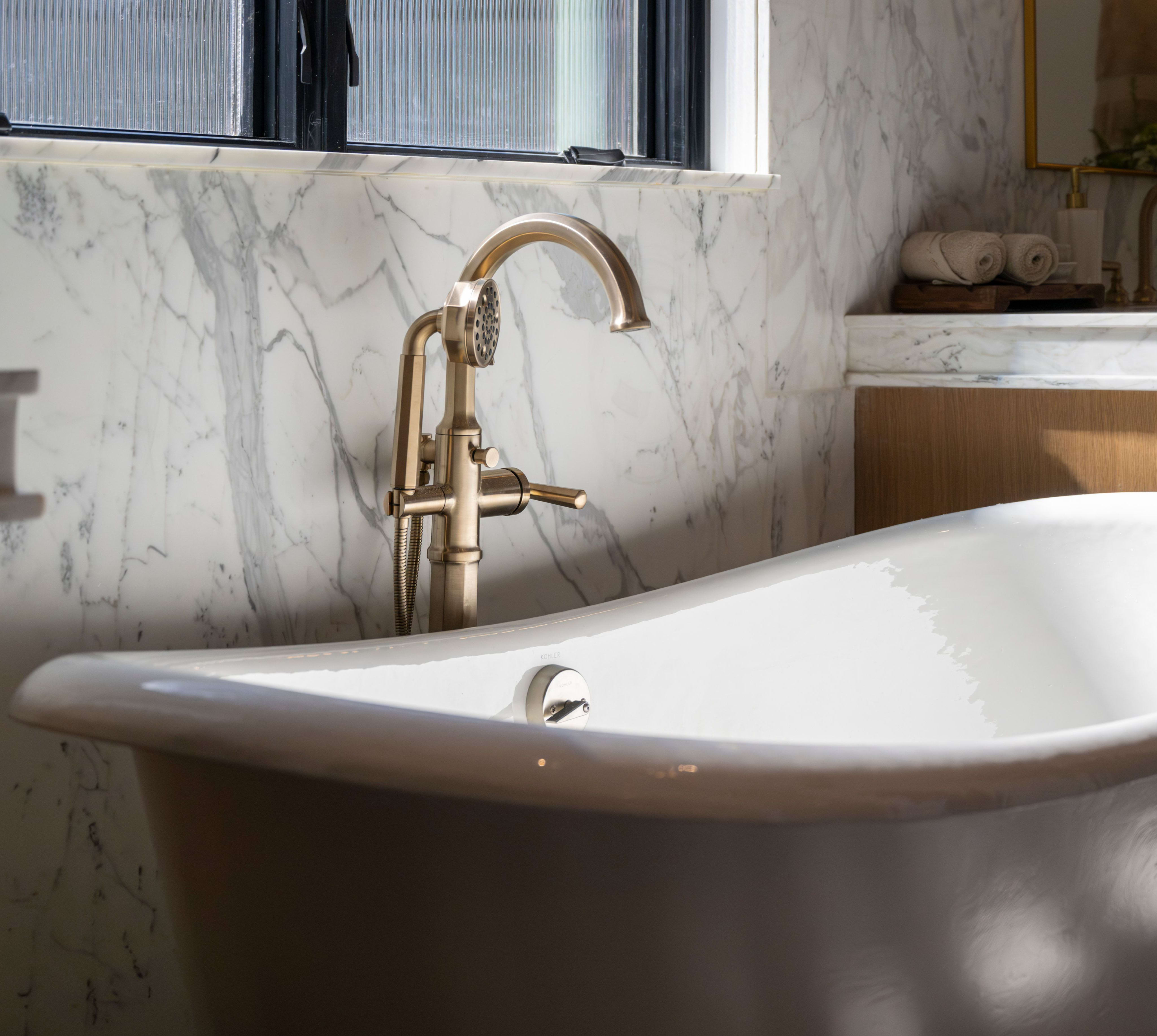 A white clawfoot bathtub with a brass faucet sits in the foreground against a marble-tiled wall, with a vanity and sink visible in the blurred background.