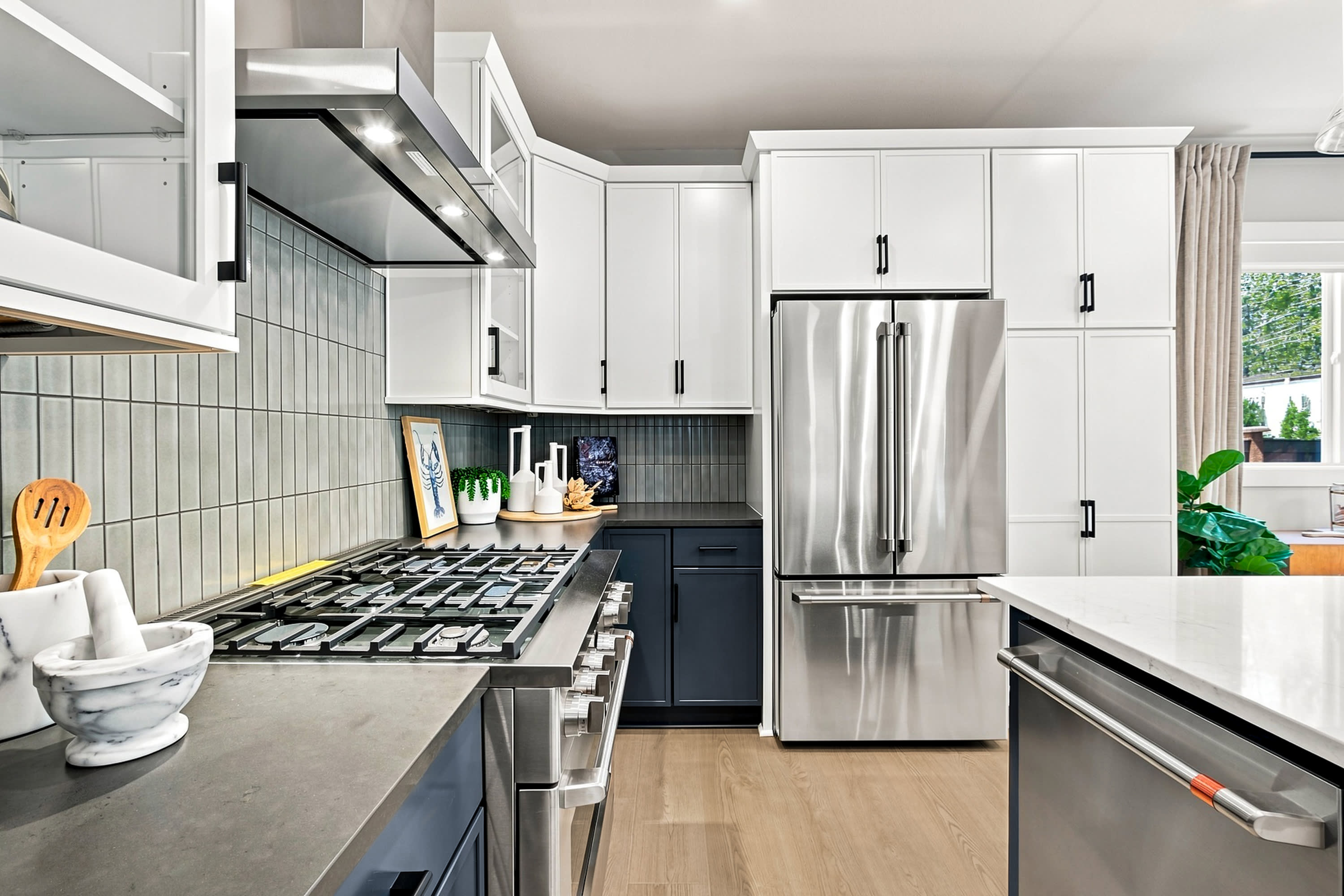 A modern kitchen features a stainless steel gas range and marble mortar in the foreground, with white cabinetry and a stainless steel refrigerator in the background.