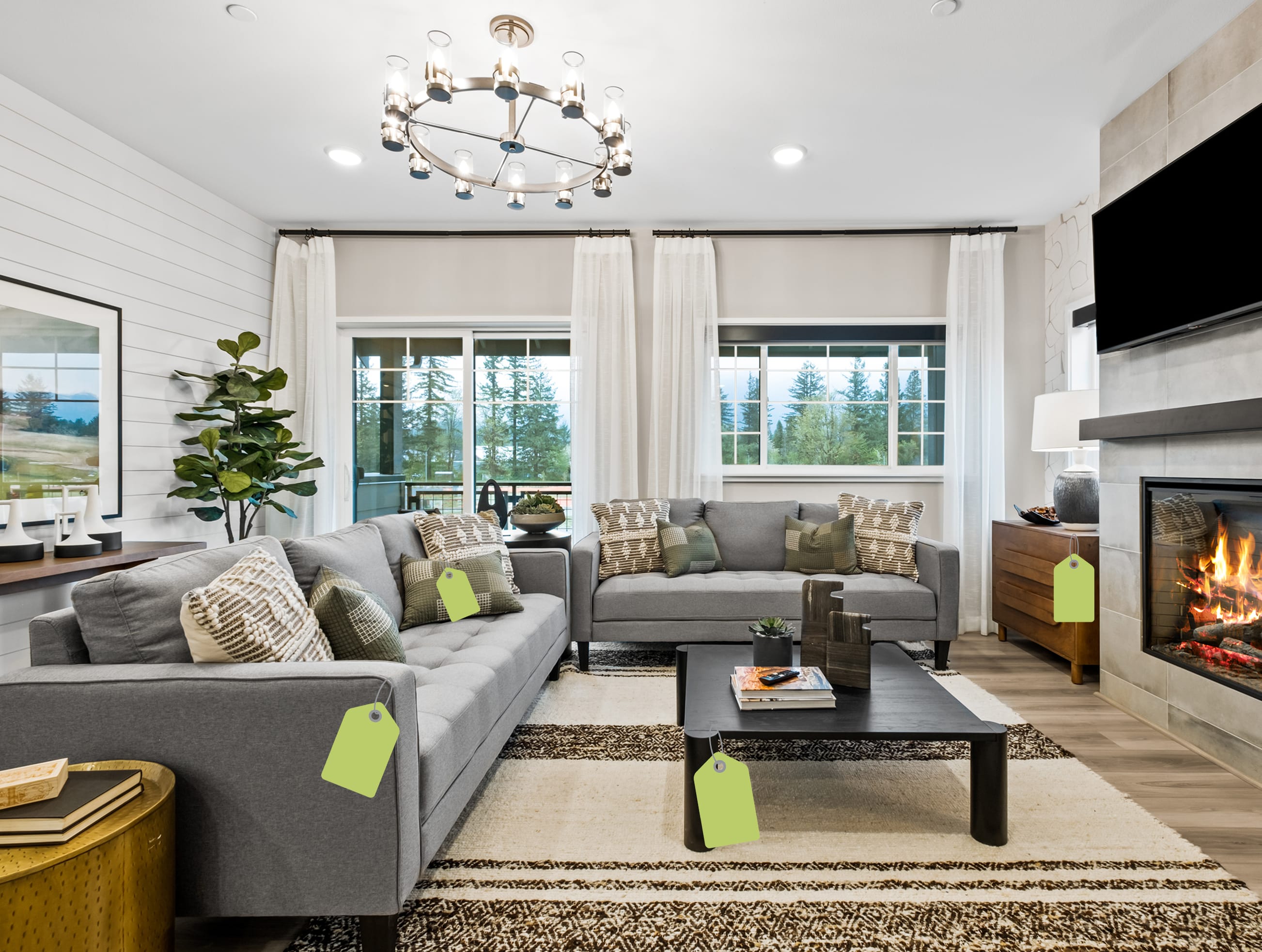 A modern living room features two grey sofas and a black coffee table with green price tags in the foreground, set against a background of white curtains, large windows, and a fireplace.