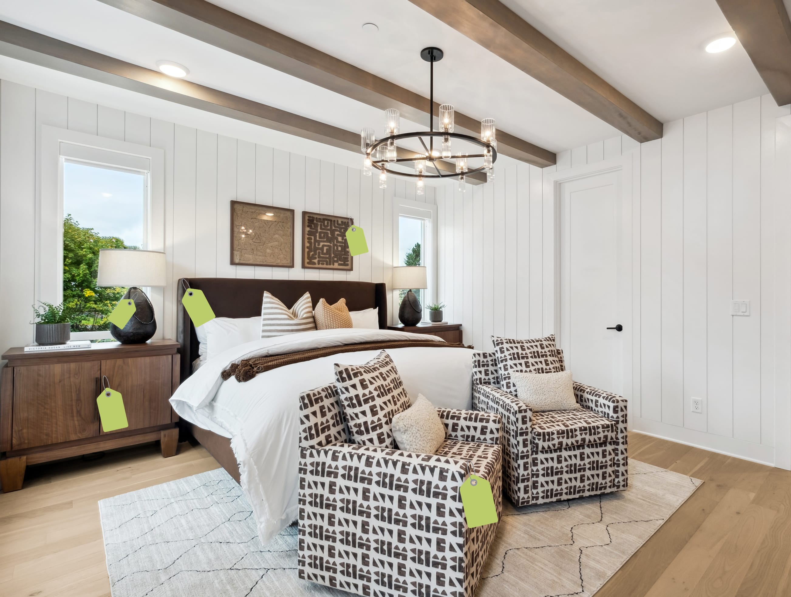 Two patterned armchairs sit on a light-colored rug in the foreground of a bright bedroom featuring a brown leather bed and white paneled walls with wooden ceiling beams in the background.