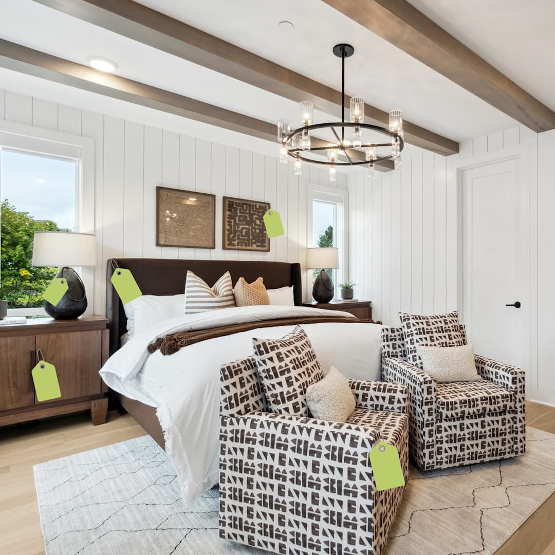 Two patterned armchairs sit in the foreground of a bright bedroom featuring a brown leather bed and white paneled walls with wooden ceiling beams.