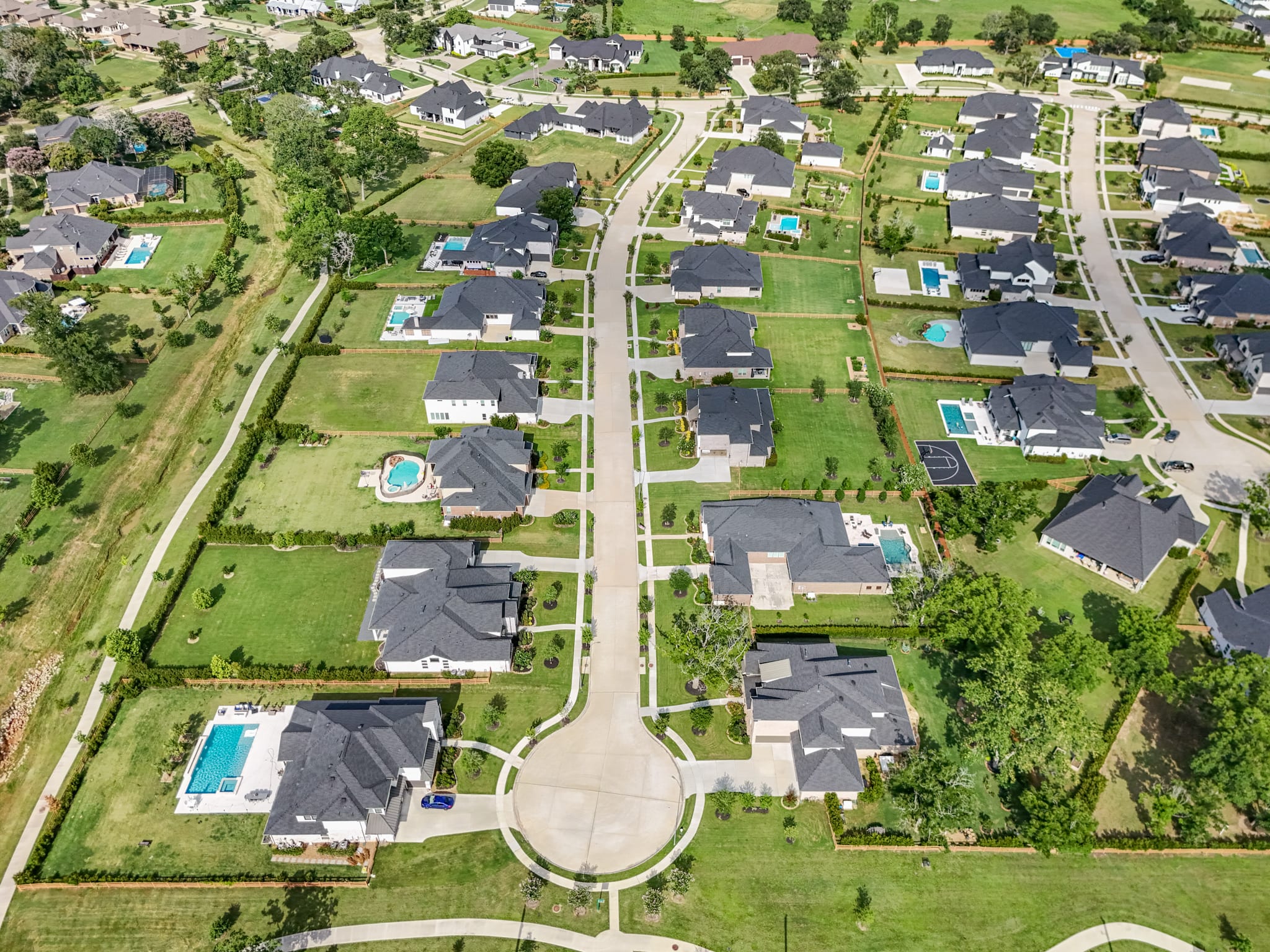 An aerial view of a residential neighborhood with neatly arranged houses, lush green lawns, and winding roads, surrounded by a picturesque countryside landscape.