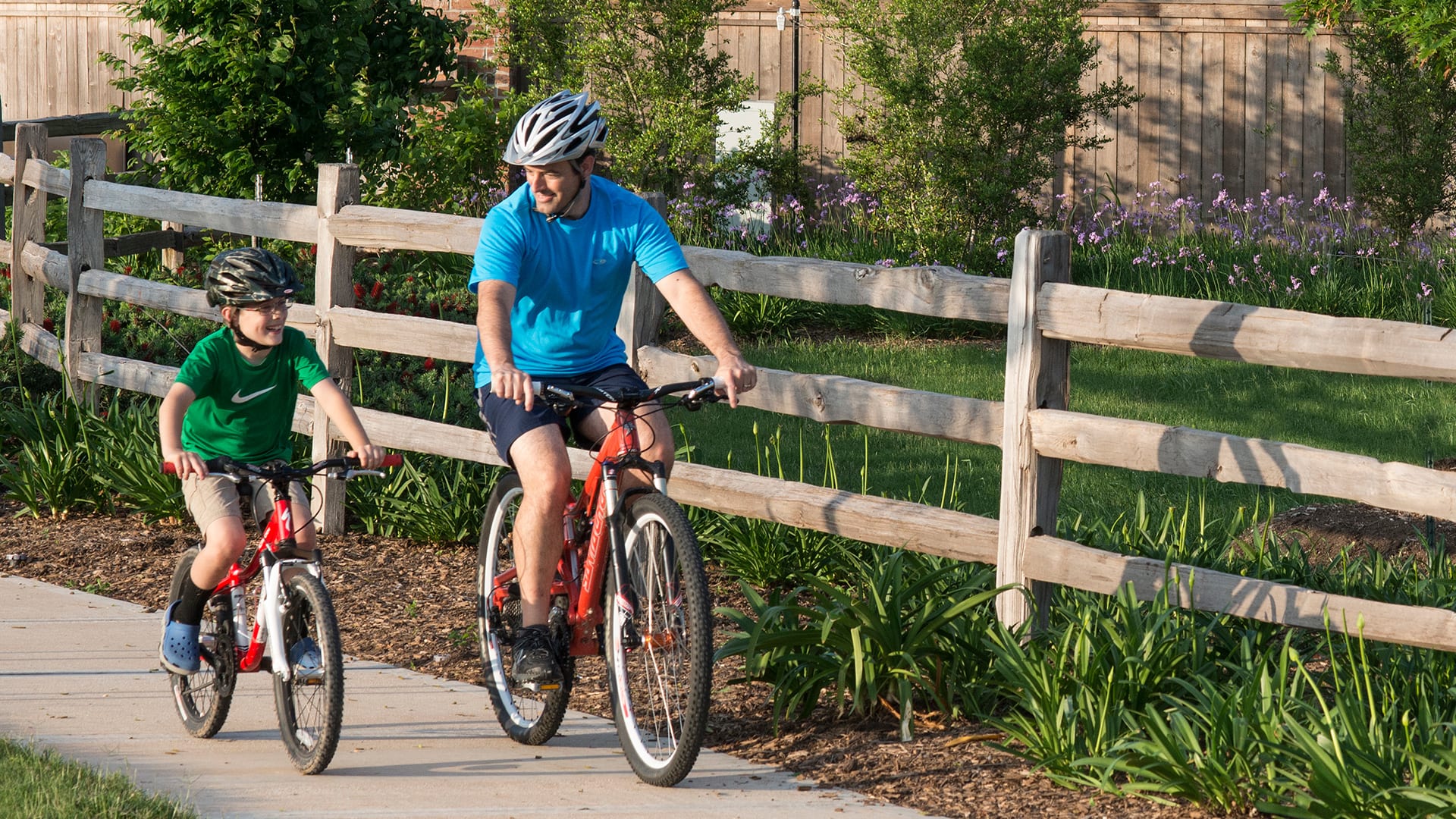 Two cyclists, one in a blue shirt and the other in a green shirt, are riding their bicycles on a path surrounded by a wooden fence and lush greenery in the background.