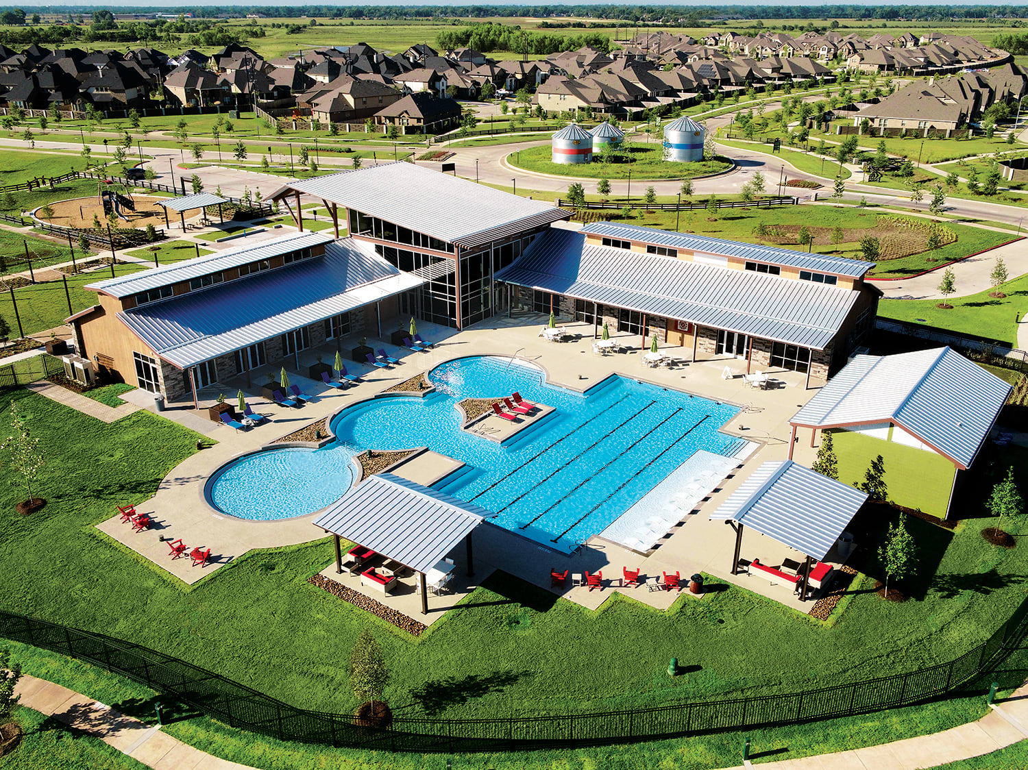 A large outdoor swimming pool complex surrounded by numerous residential buildings and lush green landscaping in a scenic rural setting.