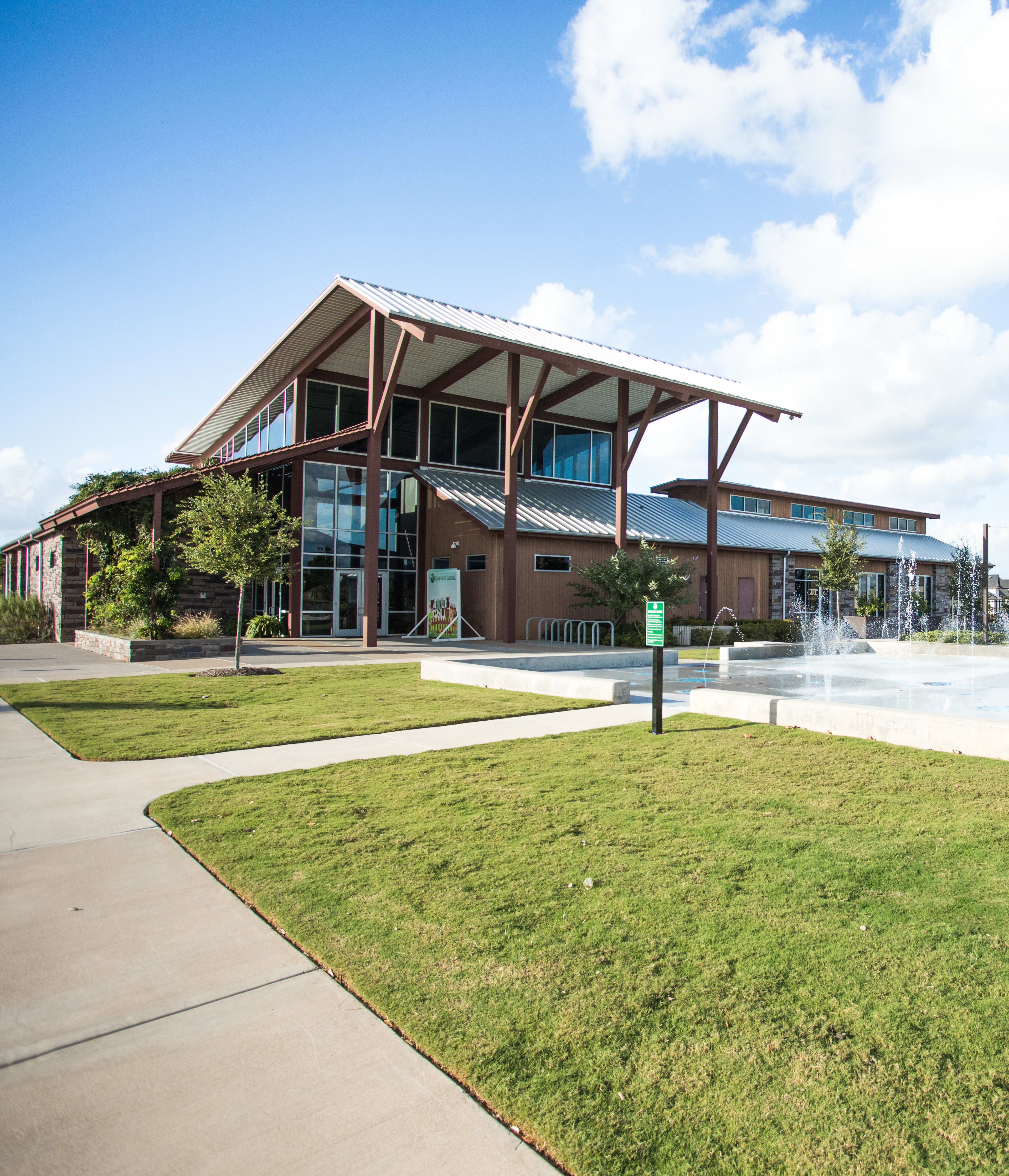 A modern, multi-story building with large windows and a covered porch stands in a grassy area with a water fountain feature in the foreground, against a backdrop of a clear blue sky with fluffy white clouds.
