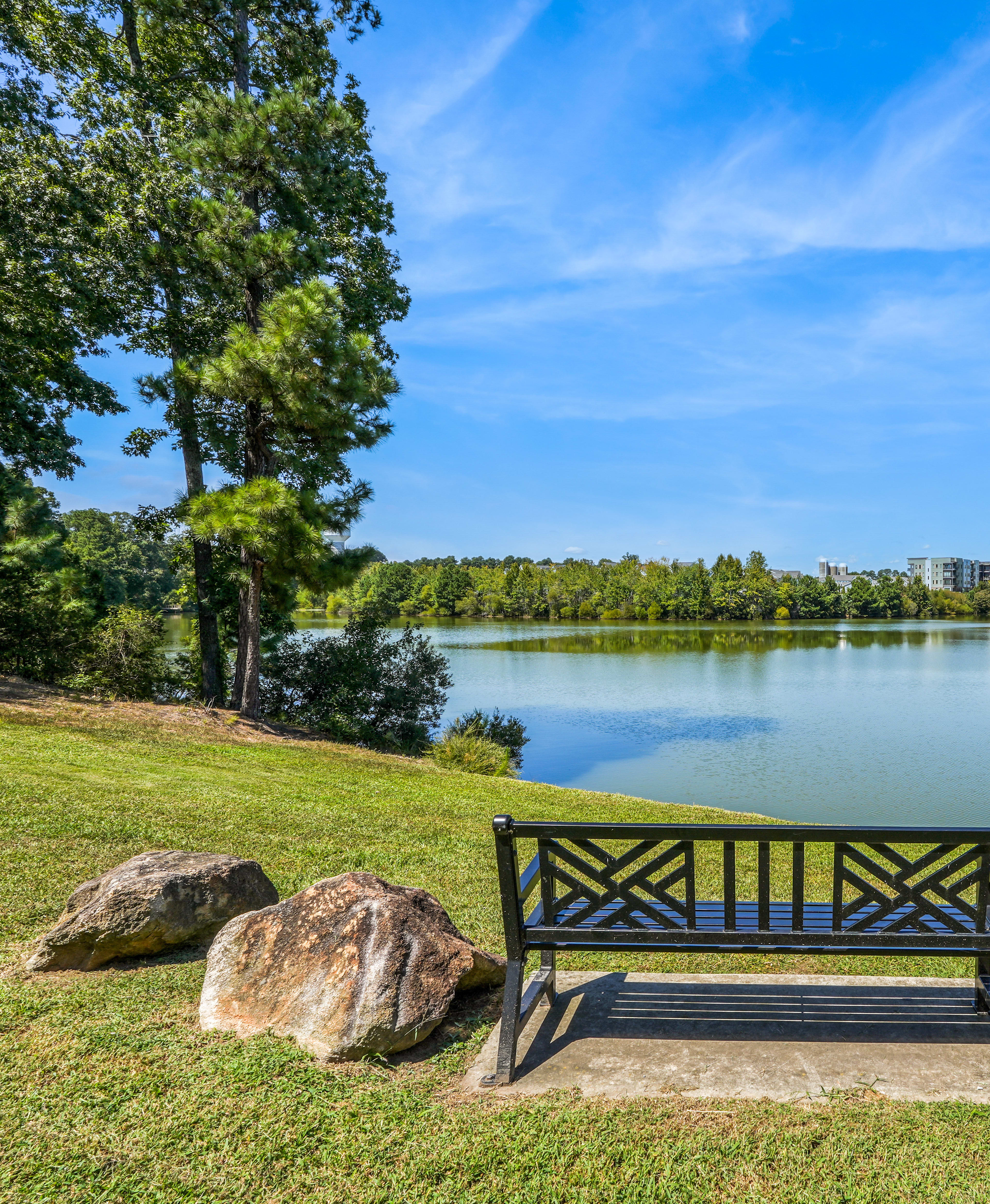 A peaceful lakeside scene with a wooden bench overlooking the calm waters, surrounded by lush greenery and a clear blue sky.