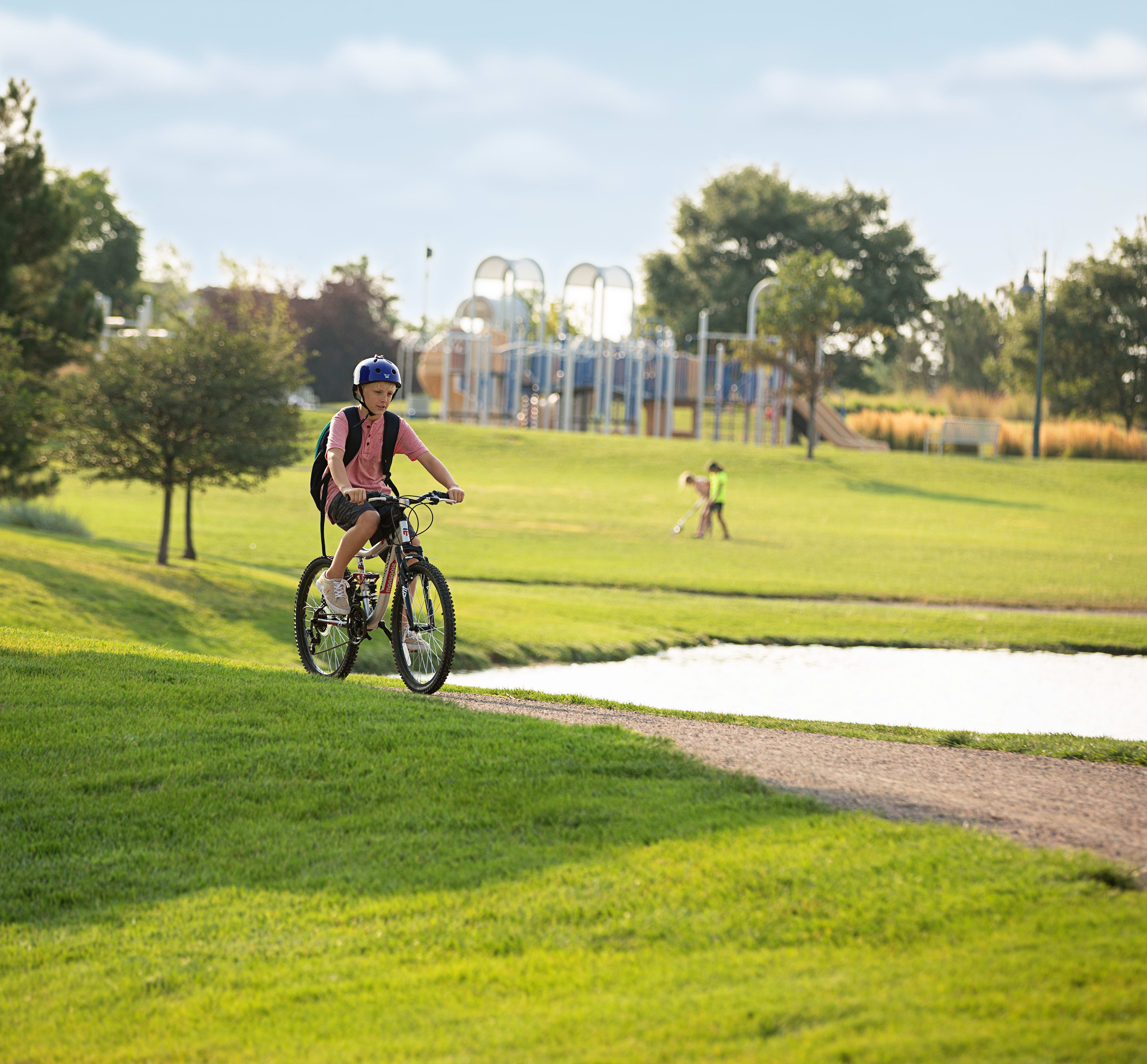 A person on a bicycle rides along a paved path through a lush, green park with trees and playground equipment visible in the background.
