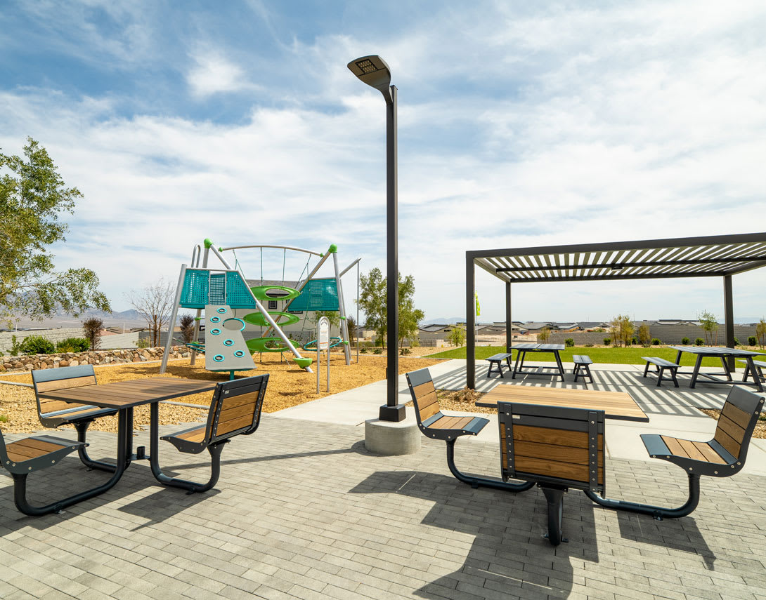 A paved outdoor seating area with picnic tables and benches, surrounded by a playground with colorful equipment in the background, under a clear blue sky with scattered clouds.