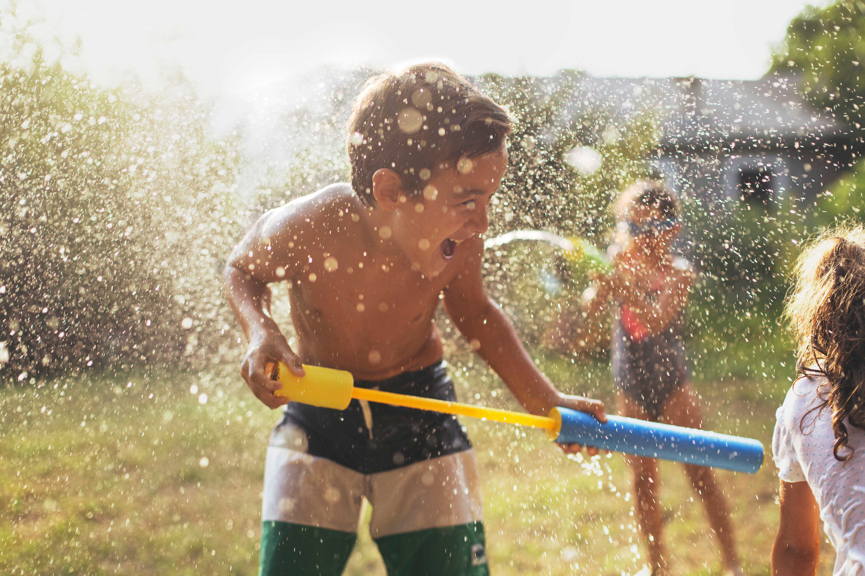 A shirtless man is holding a water gun and spraying water on a group of people in a sunny, outdoor setting with lush greenery in the background.