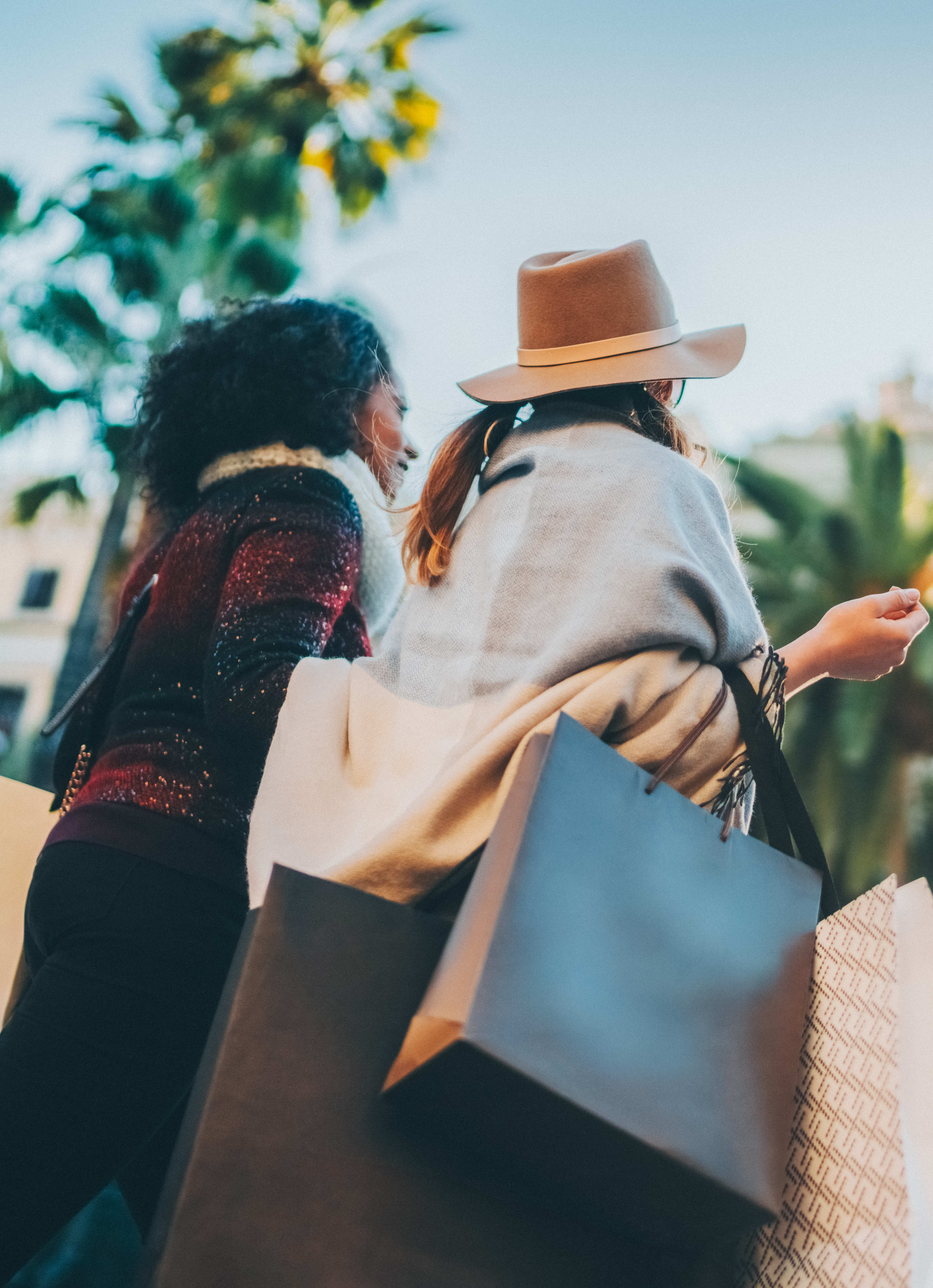 Two people, a man and a woman, are walking together while carrying shopping bags in a sunny, palm tree-lined street.