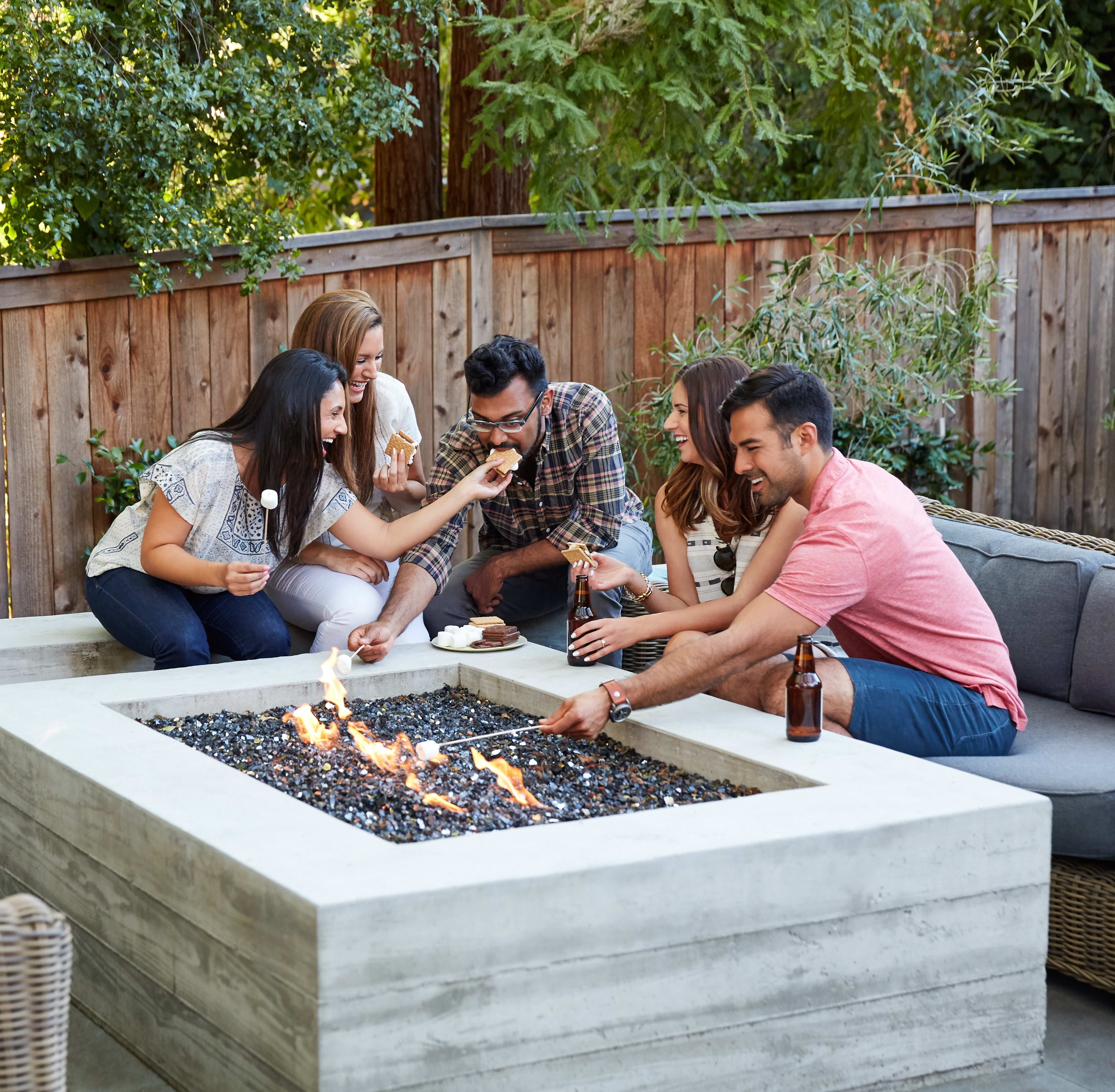 A group of friends gathered around a concrete fire pit, enjoying each other's company in a cozy outdoor setting surrounded by lush greenery and a wooden fence.