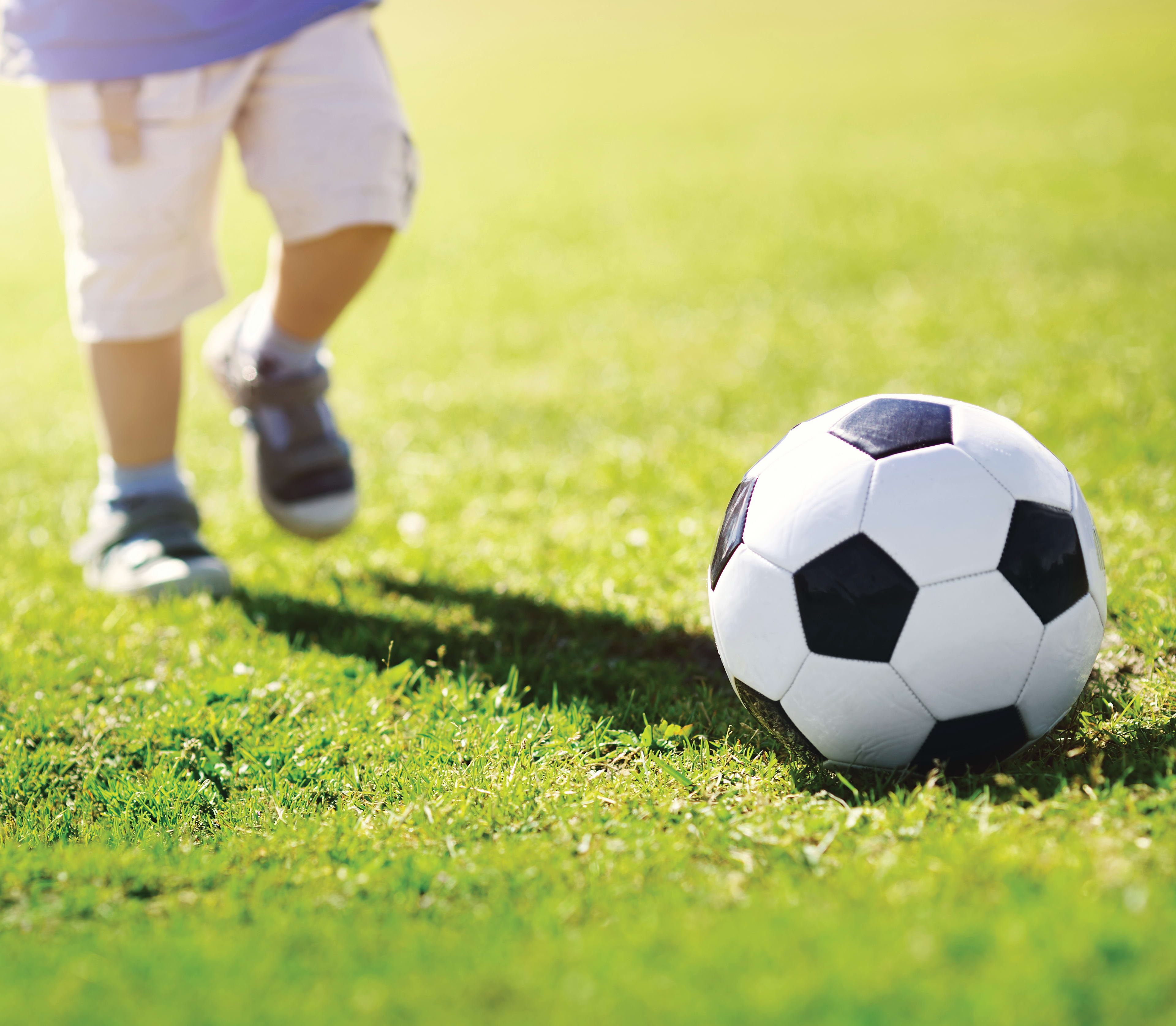 A soccer ball rests on a lush, green field, with a person's legs visible in the foreground, suggesting they are about to kick the ball.