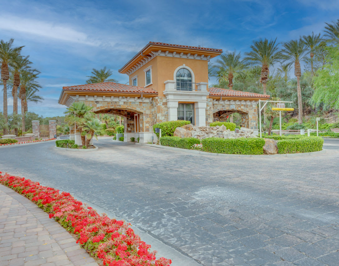 A grand, Mediterranean-style villa with arched windows and a tiled roof sits amidst a lush, landscaped garden featuring vibrant red flowers lining the paved driveway leading to the entrance.