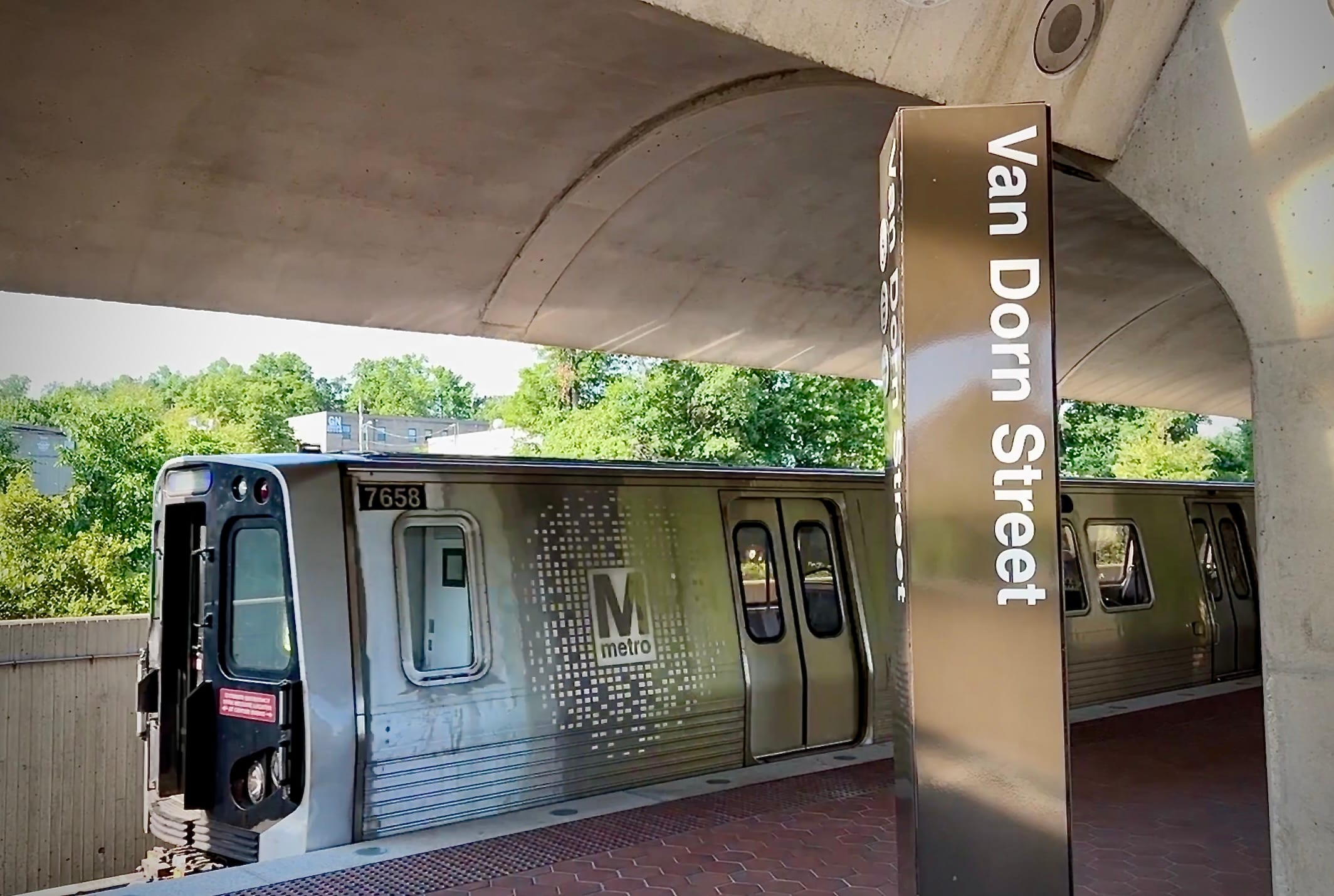 A subway train with the "Van Dorn Street" sign is parked under a concrete overpass, surrounded by greenery in the background.