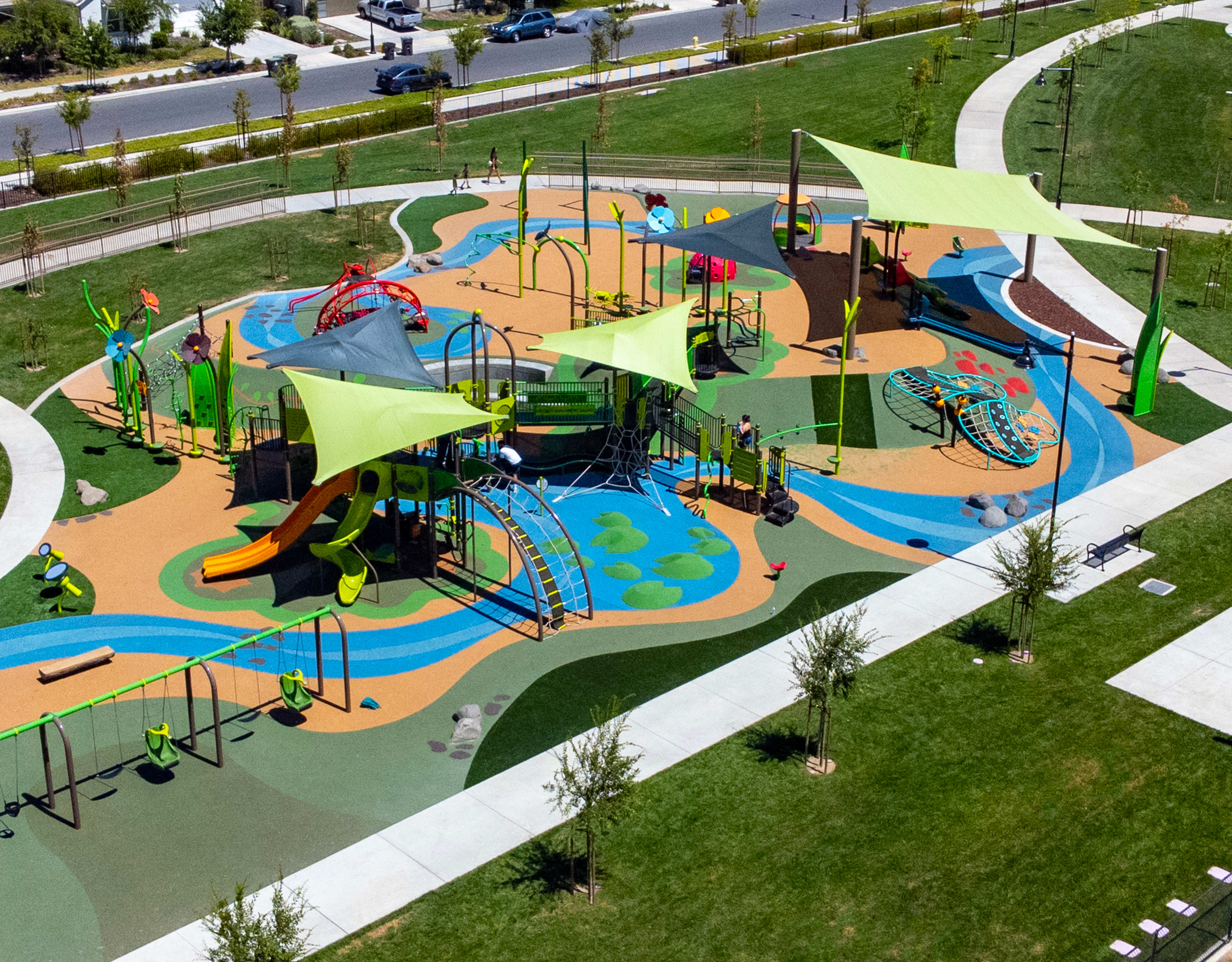A vibrant and colorful playground with various play structures, including slides, climbing equipment, and a splash pad, surrounded by lush greenery and a paved walkway.