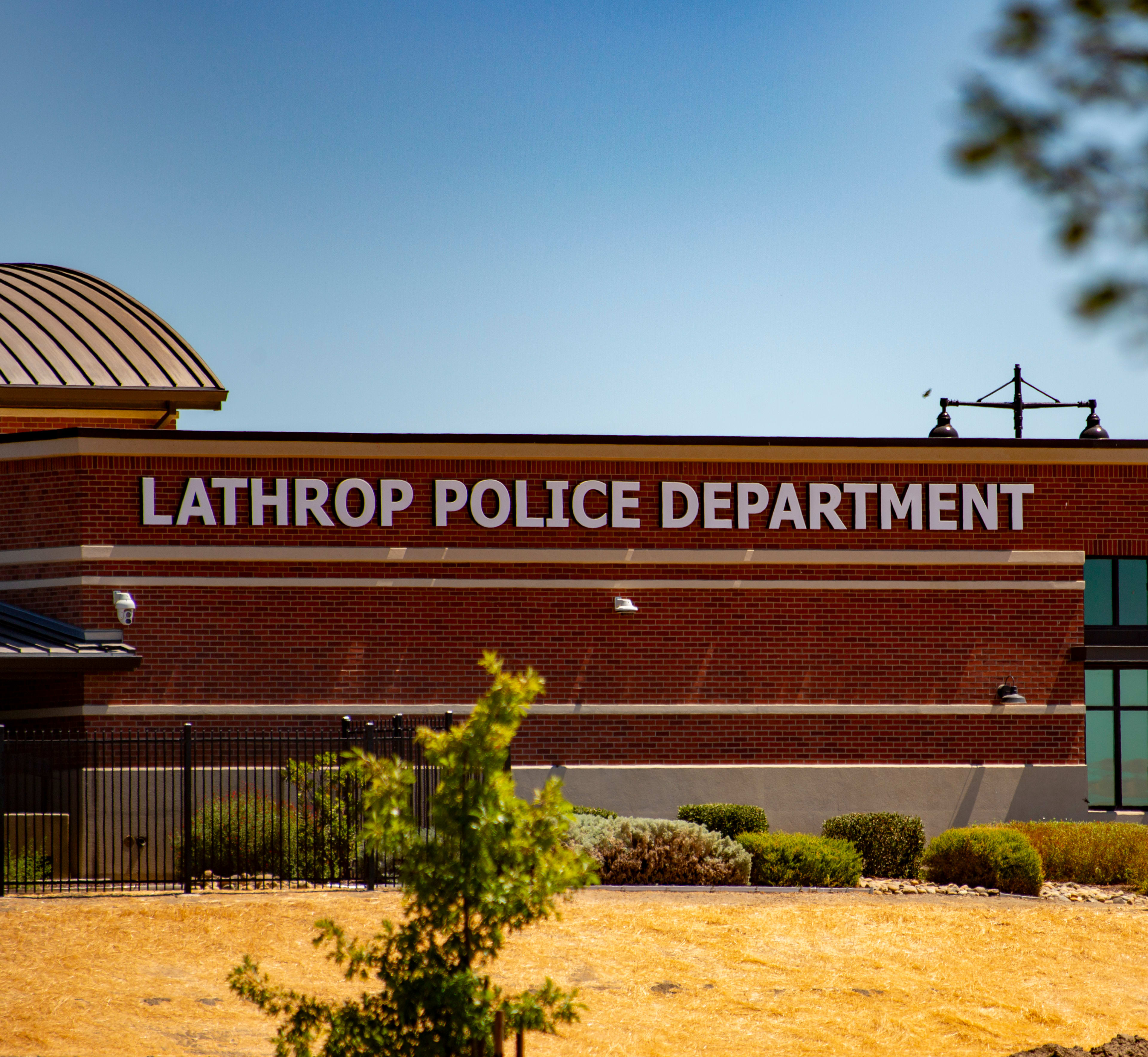 A brick building with the words "Lathrop Police Department" prominently displayed, surrounded by a sandy landscape and trees in the background.