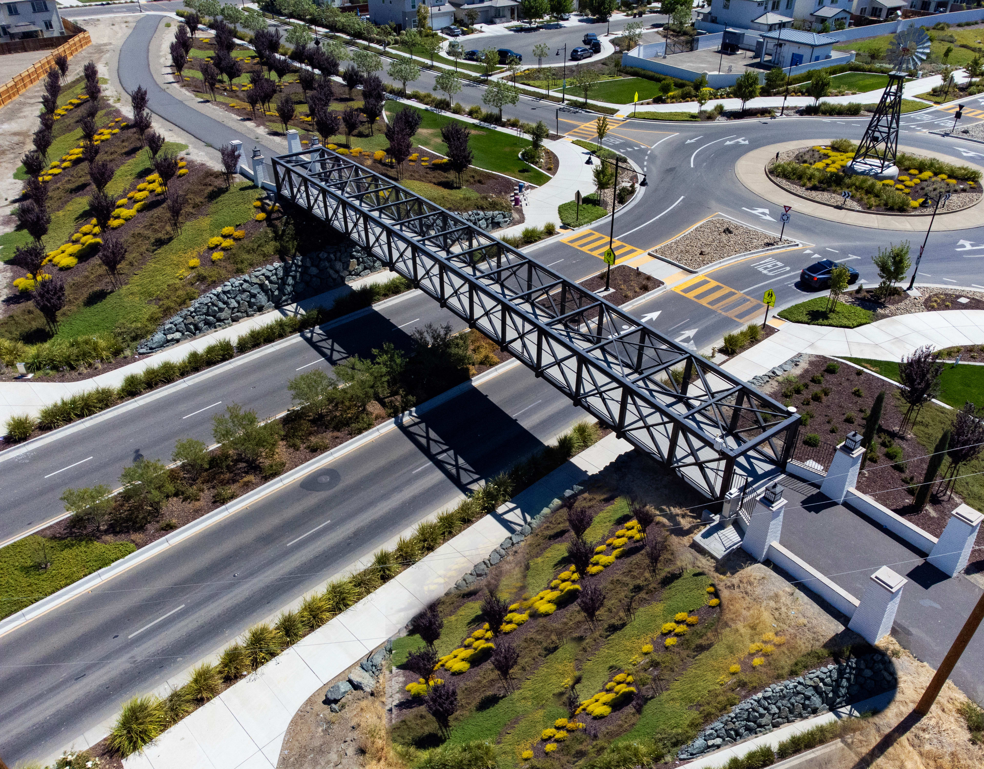 A large metal bridge spans across a road, surrounded by a landscaped area with trees and flowers in the foreground, and residential buildings in the background.