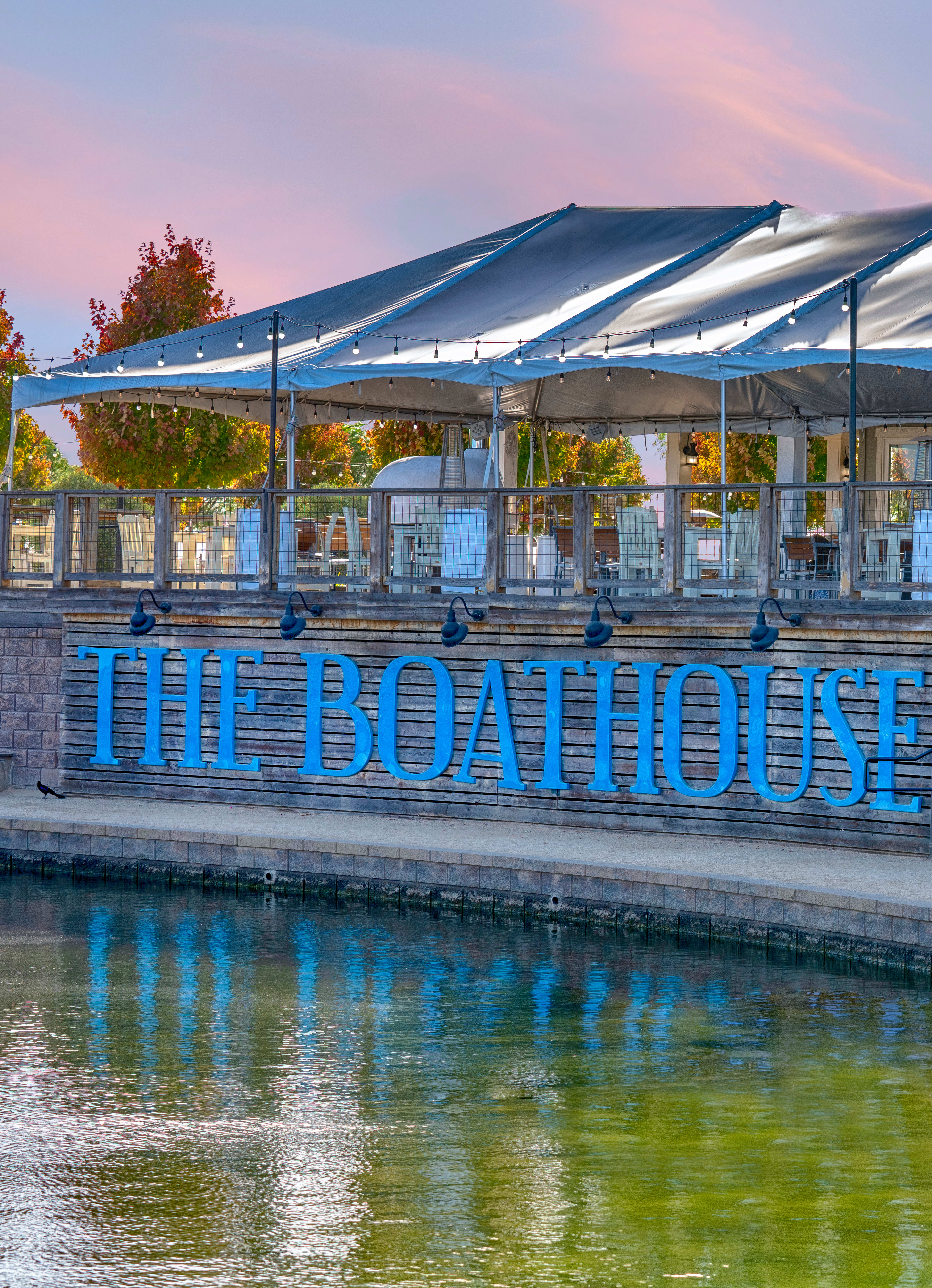 A modern boathouse structure with a curved roof and glass walls stands beside a calm body of water, surrounded by trees and a colorful sky at sunset.