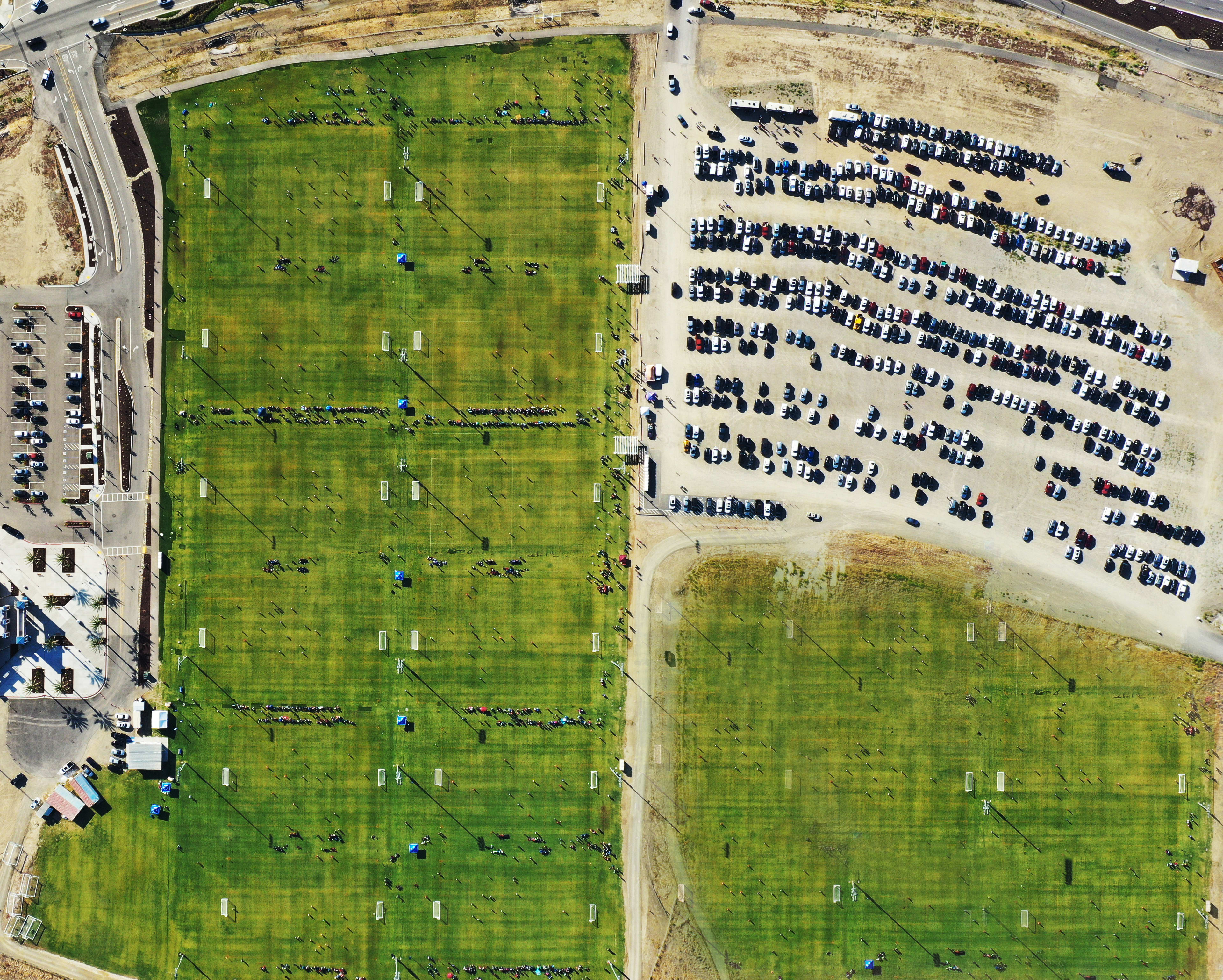 An aerial view of a baseball stadium surrounded by parking lots and green fields, with people visible on the field and in the stands.