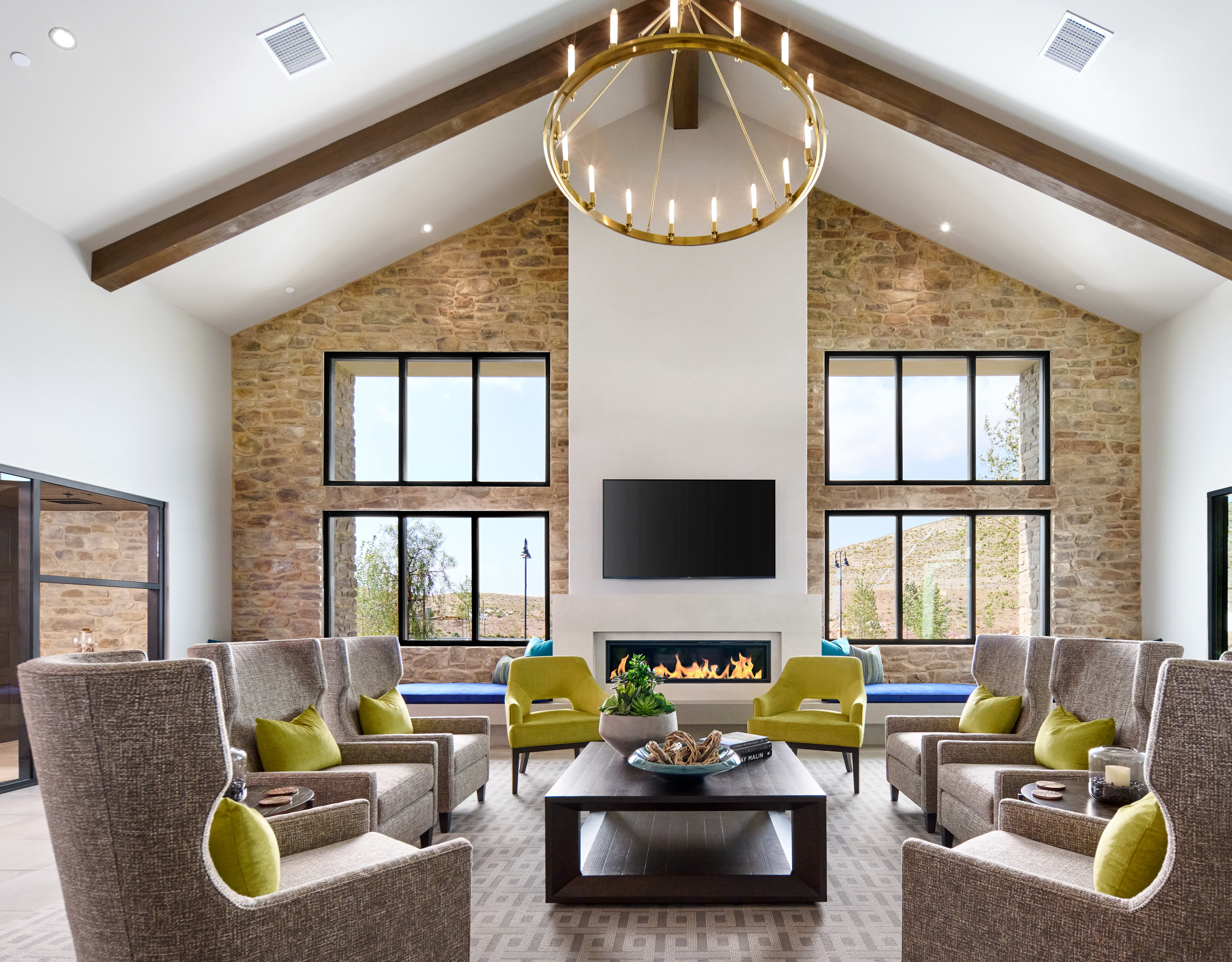 A spacious and modern living room with high ceilings, exposed brick walls, and a large fireplace surrounded by comfortable seating arrangements, including gray sofas and green accent chairs, all centered around a wooden coffee table.