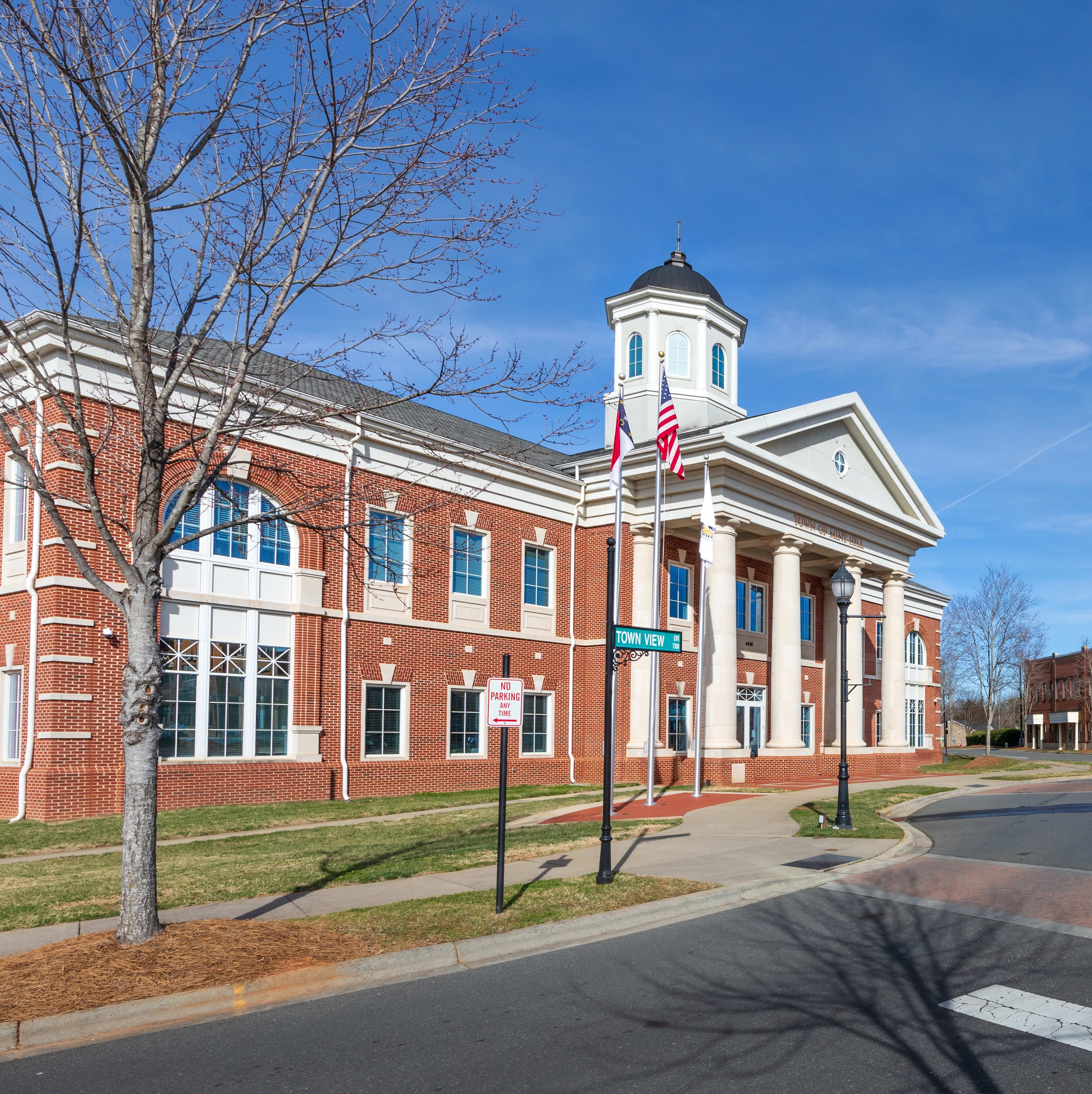 A brick building with a clock tower stands prominently in the foreground, surrounded by trees and a paved walkway leading to other buildings in the background.