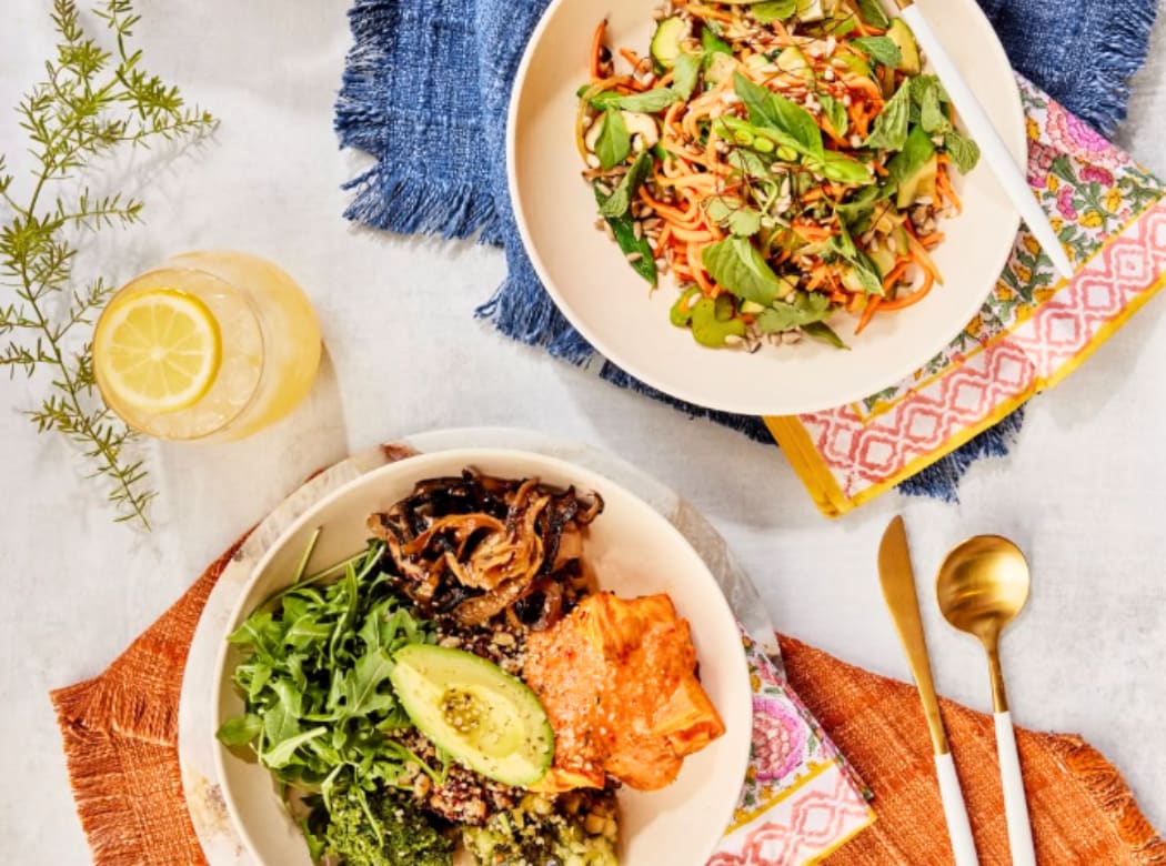 A colorful and vibrant meal featuring a variety of dishes, including a salad with fresh greens and vegetables, and a plate with rice, beans, and what appears to be a protein-rich dish. The background includes a blue and white patterned cloth, a lemon wedge, and some herbs.